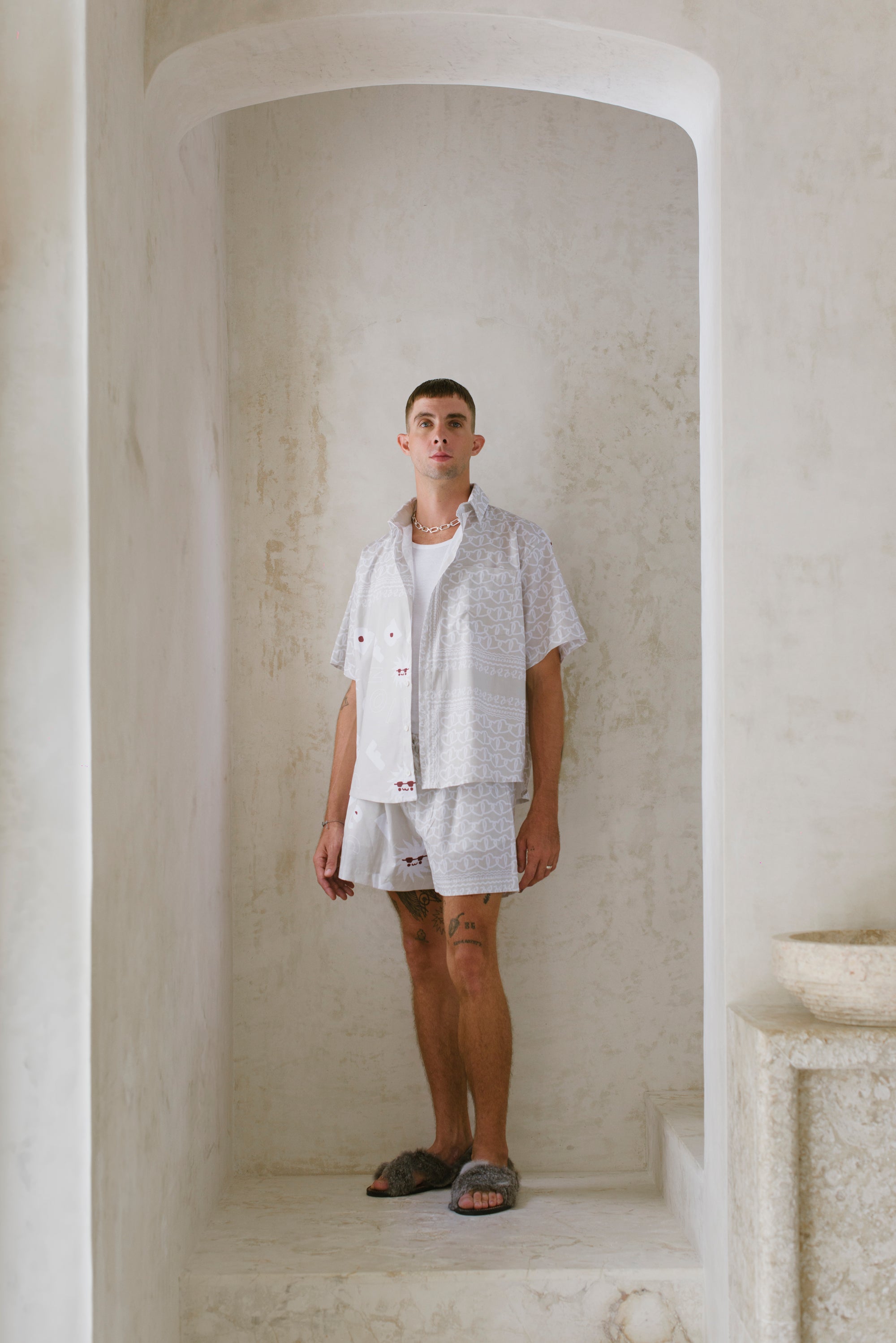Person wearing a light-colored patterned outfit standing in a minimalistic room with white walls.