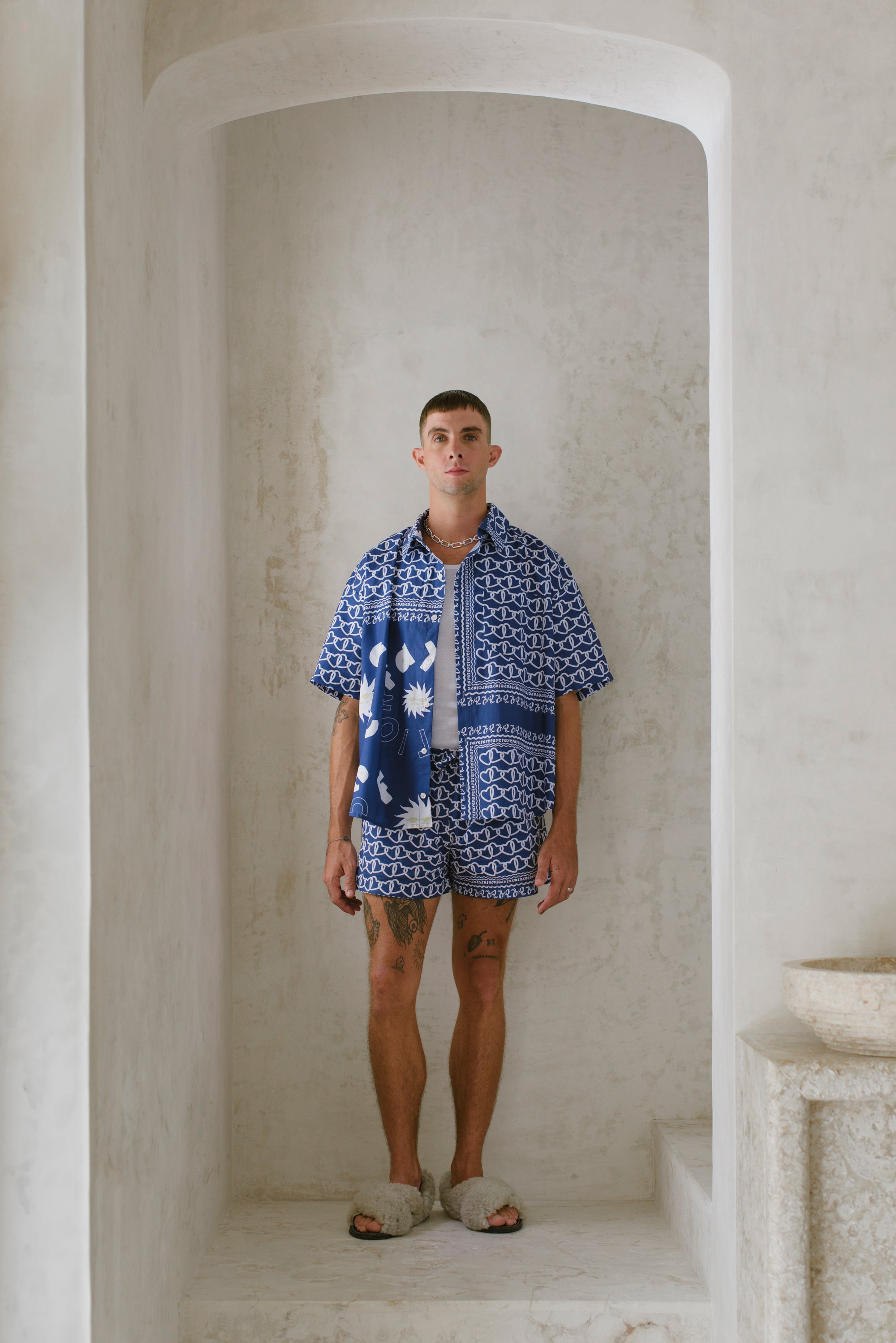 Man wearing a blue patterned shirt and shorts standing in a stone archway.