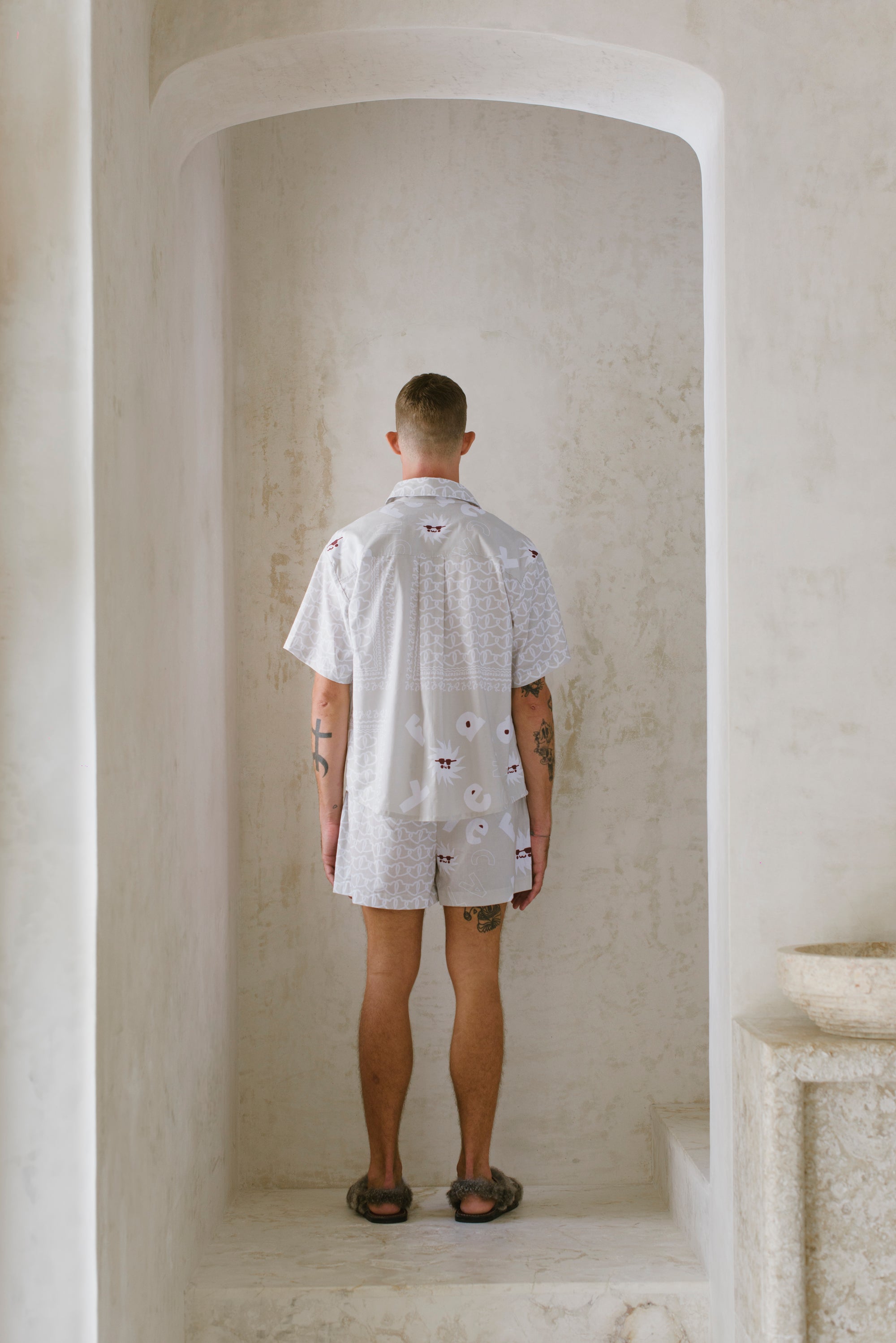 Man standing in a stone archway wearing a light-colored patterned shirt and shorts.