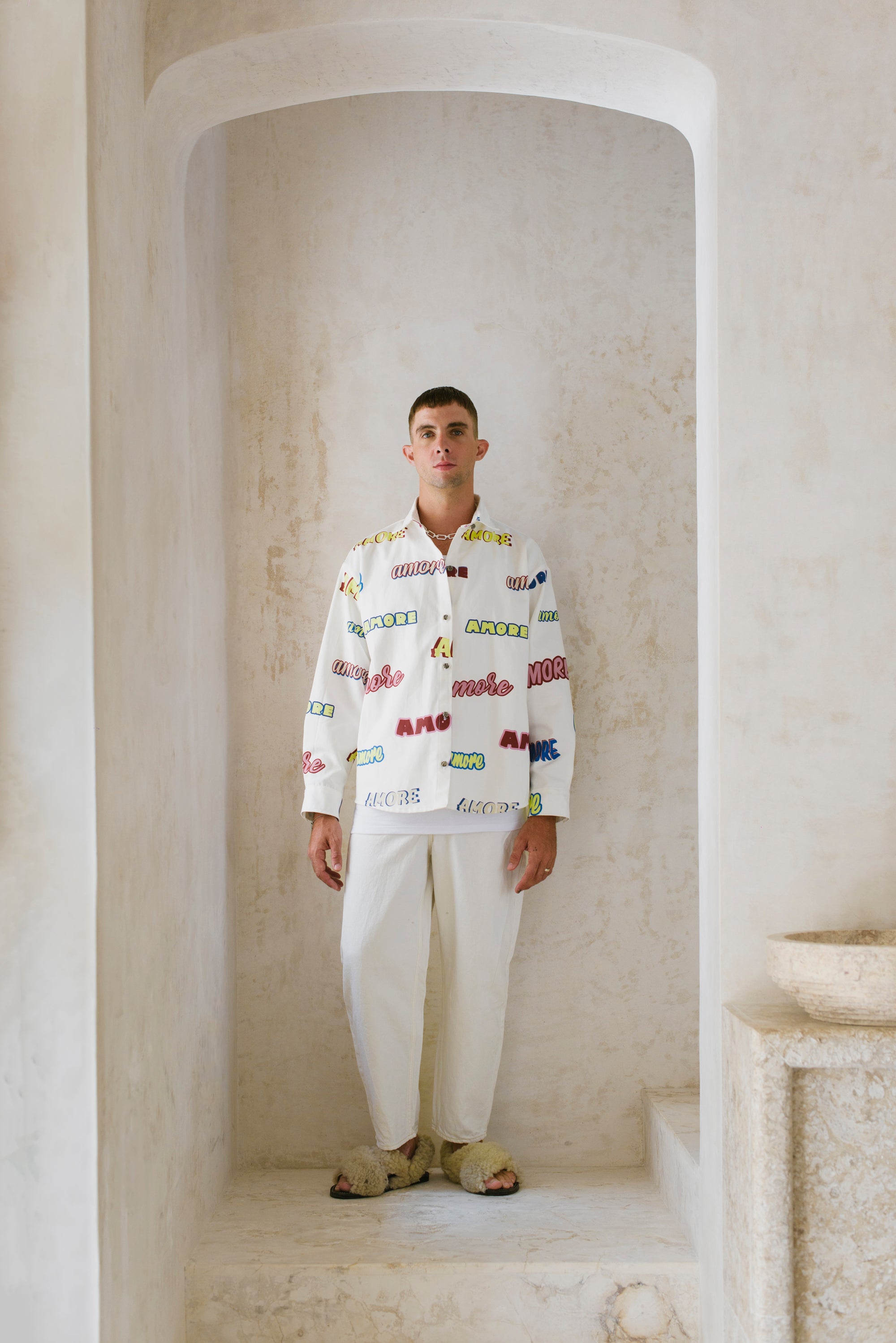 Man wearing a colorful shirt and white pants standing in a stone archway.
