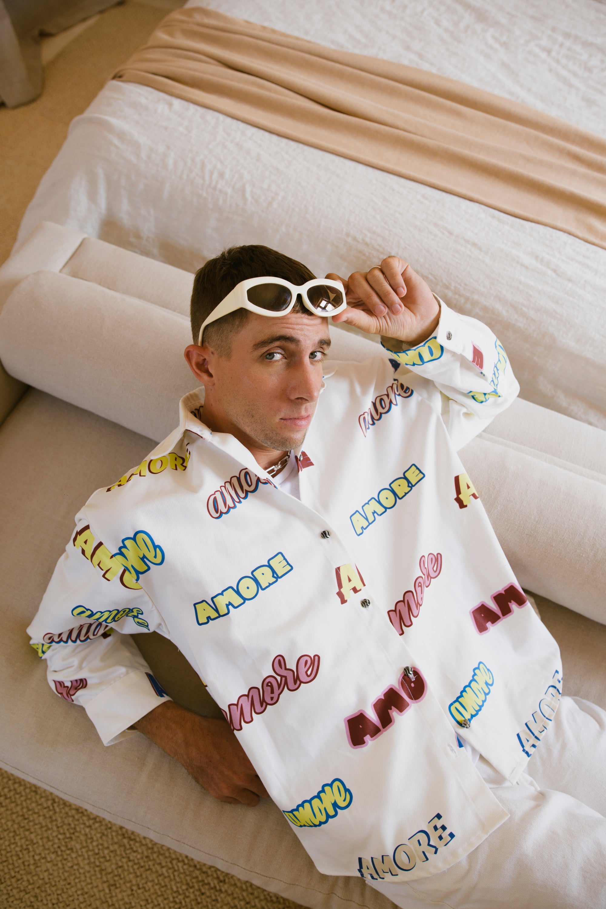 Man wearing a white overshirt with colorful text, sitting on a couch.
