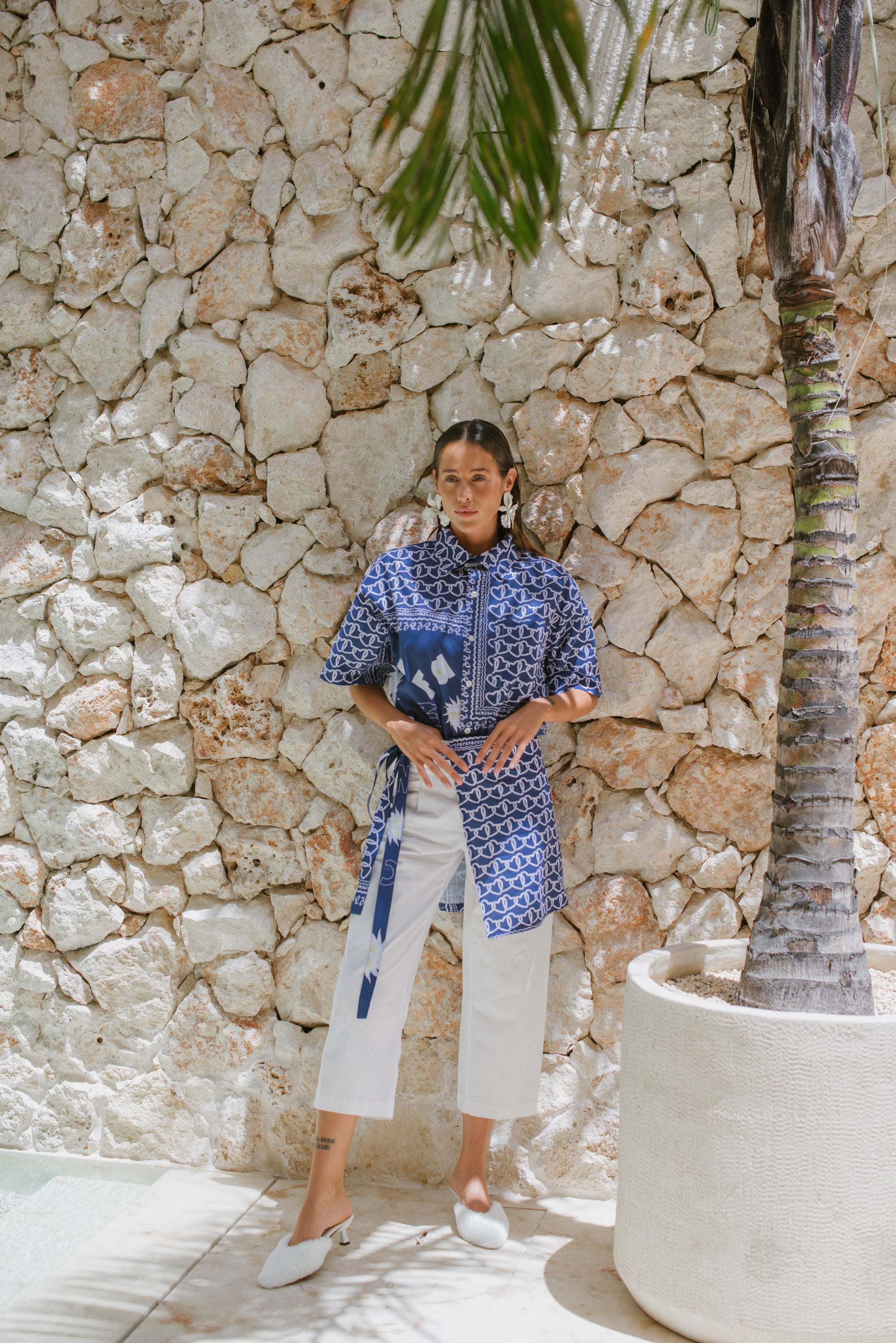 Woman in a blue patterned shirt and white pants standing against a stone wall with a palm tree.