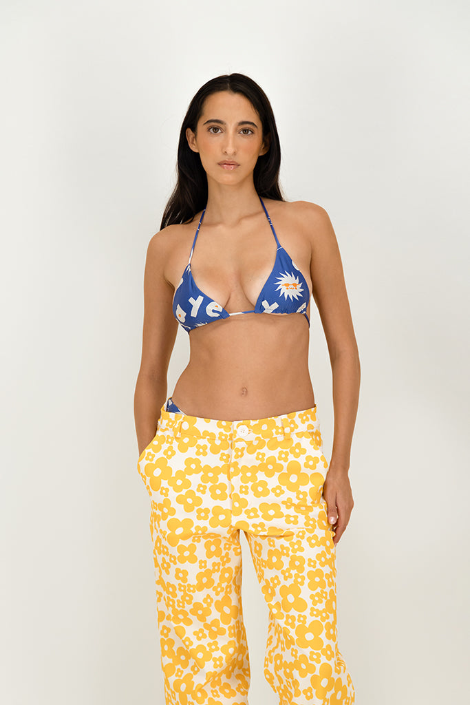 A woman modeling a two-piece swimsuit featuring a blue triangle bikini top with a white sun and letter print, paired with yellow-and-white floral print pants, against a plain light gray studio background.