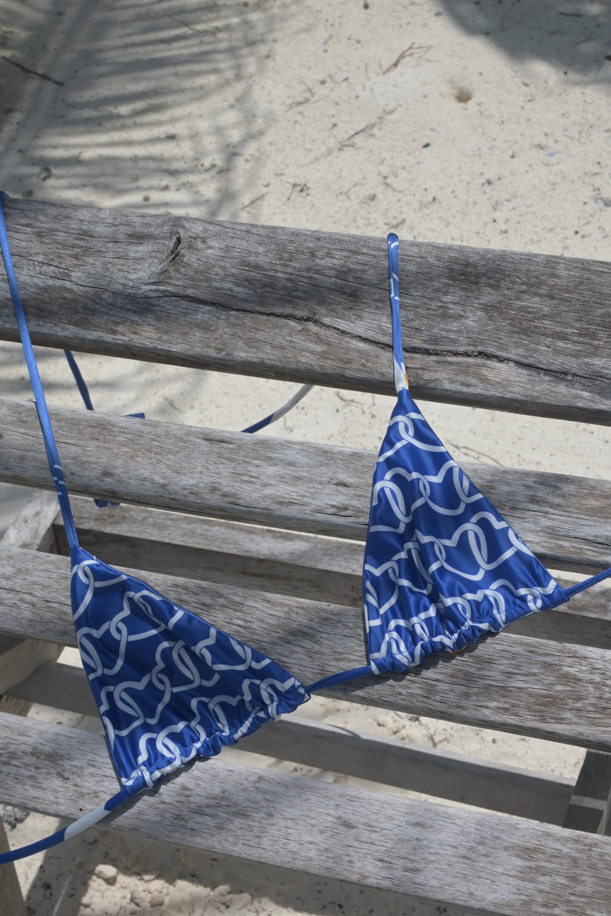 A blue and white patterned triangle bikini top hanging on a weathered wooden bench outdoors on sand.