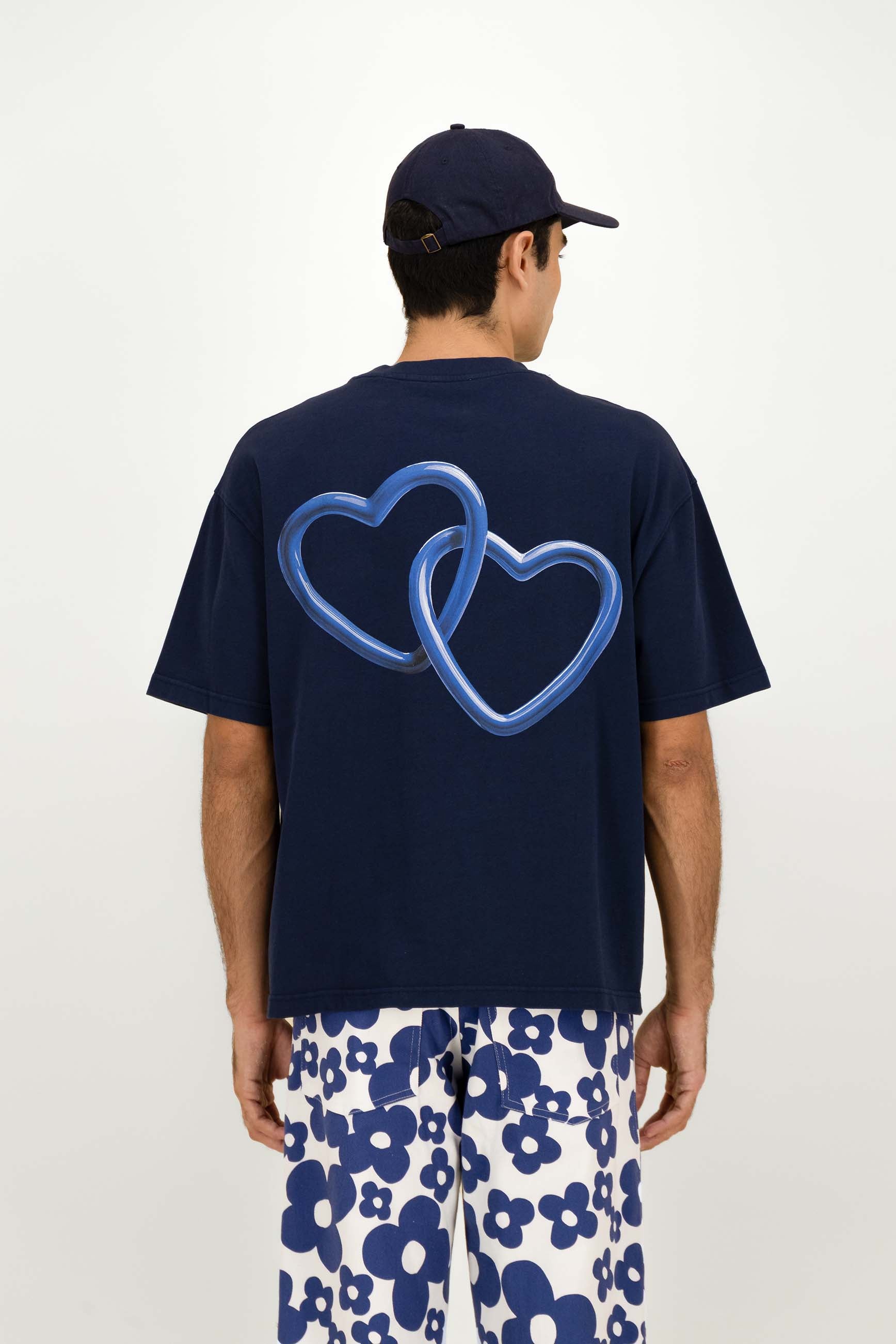 A man viewed from the back wearing a navy blue t-shirt with a large blue outlined double-heart graphic on the back, paired with white pants featuring a blue floral pattern and a navy baseball cap.