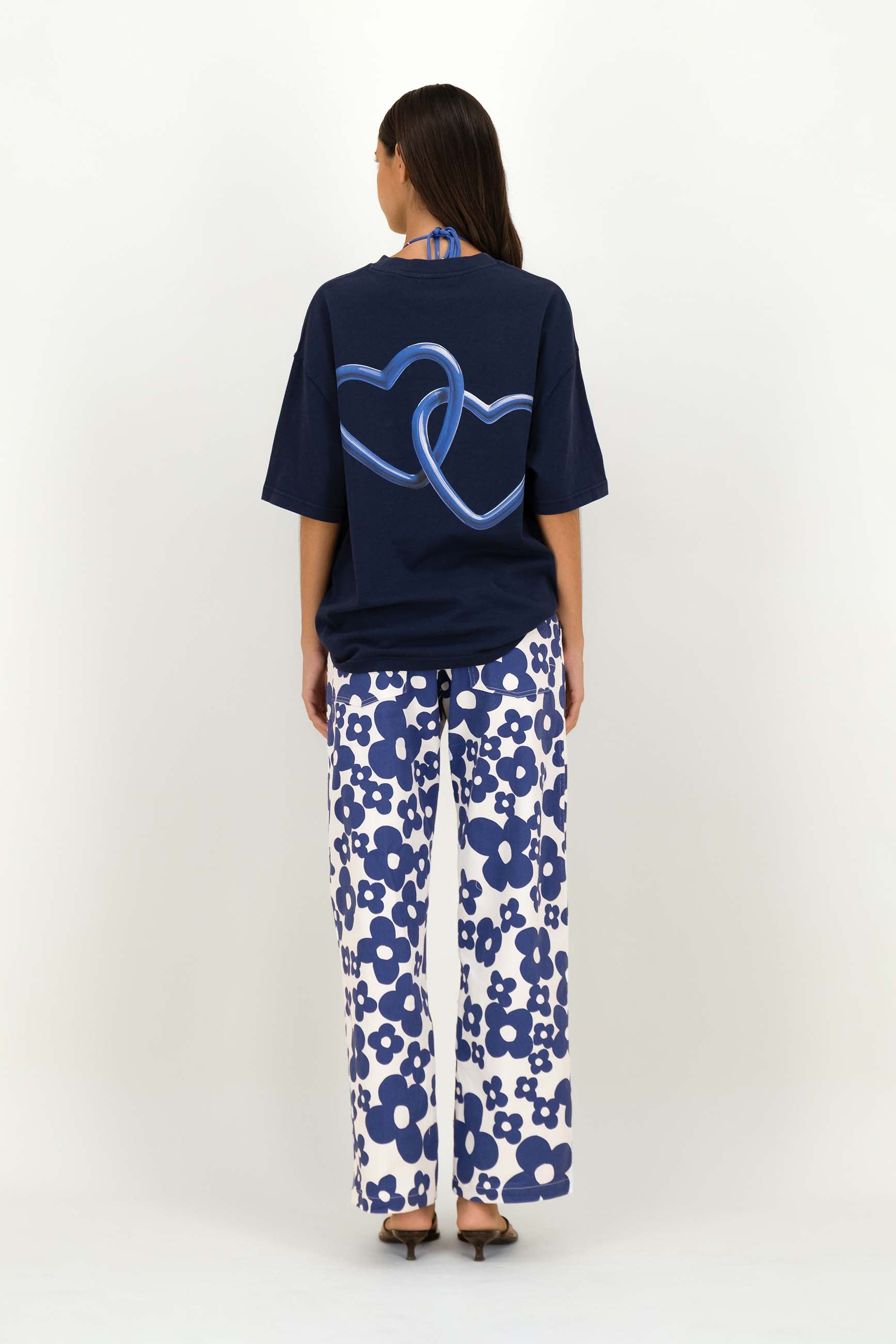 A woman shown from the back wearing a navy blue t-shirt with a blue heart graphic and white pants with a blue floral pattern, set against a plain white studio background.