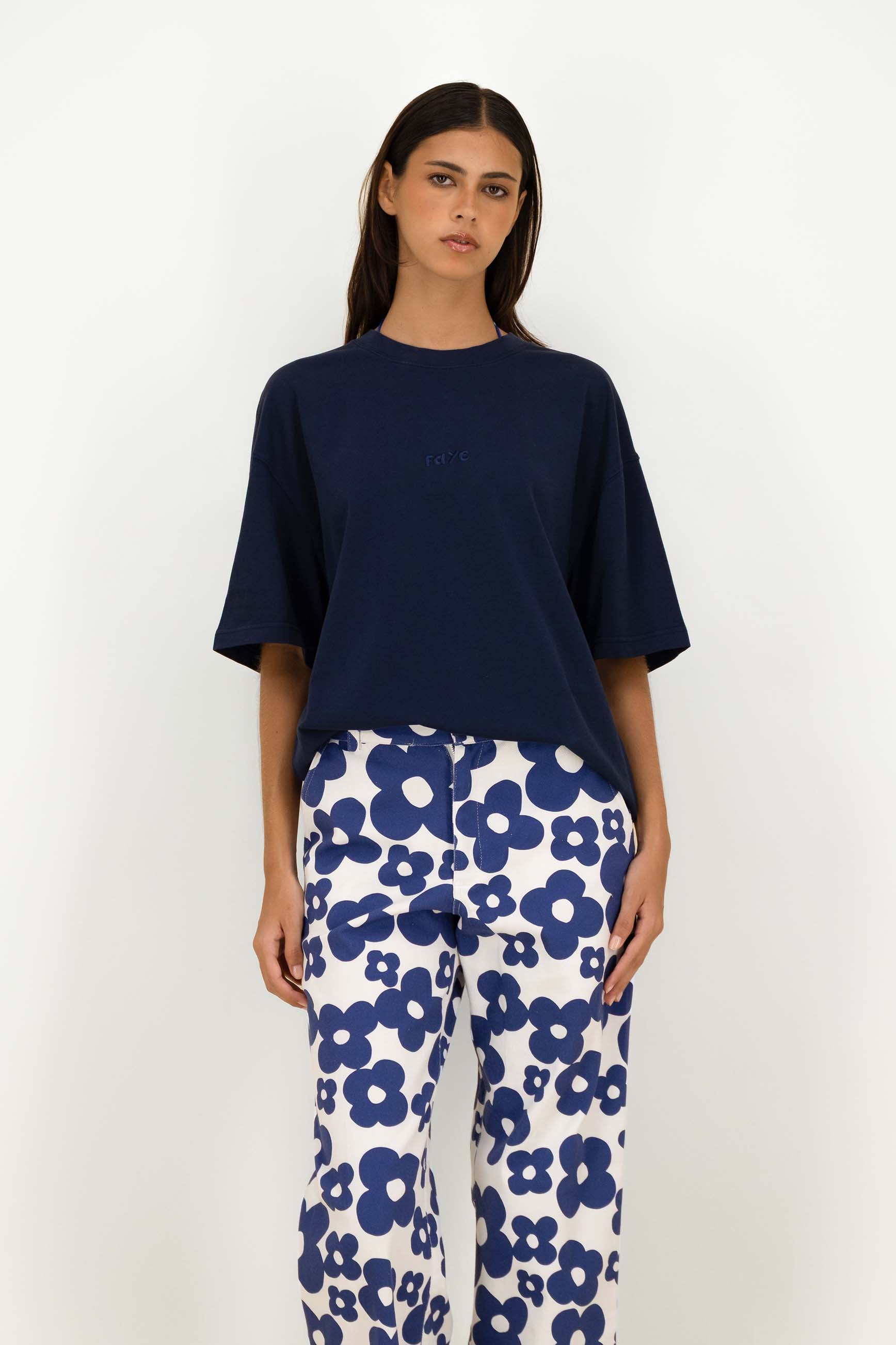 A woman modeling a navy blue short-sleeve crewneck t-shirt and white pants with a bold blue floral pattern, standing against a plain white studio background.