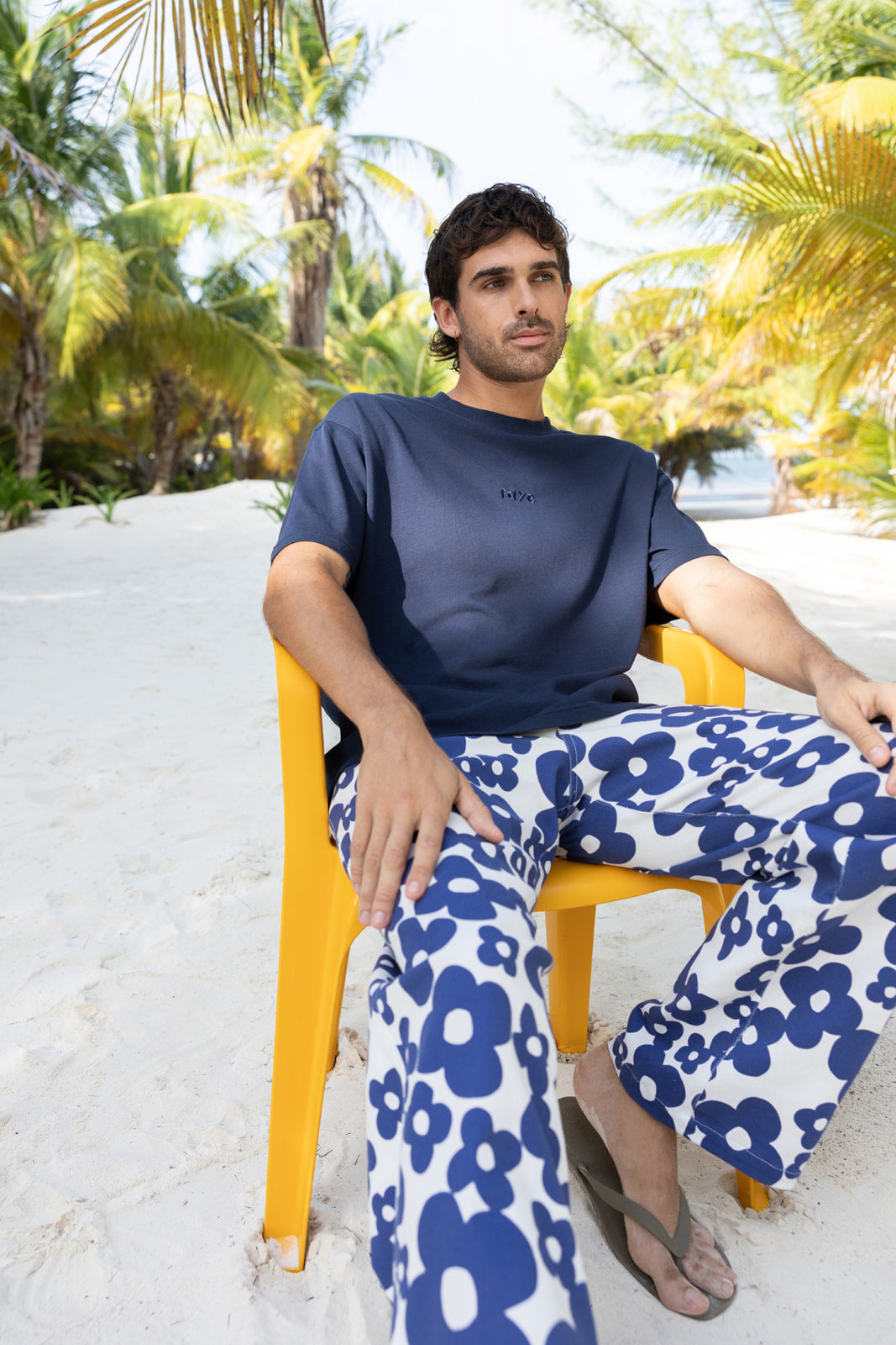 A man sitting on a yellow plastic chair on a sandy beach wearing a navy blue t-shirt and matching navy and white floral pants.