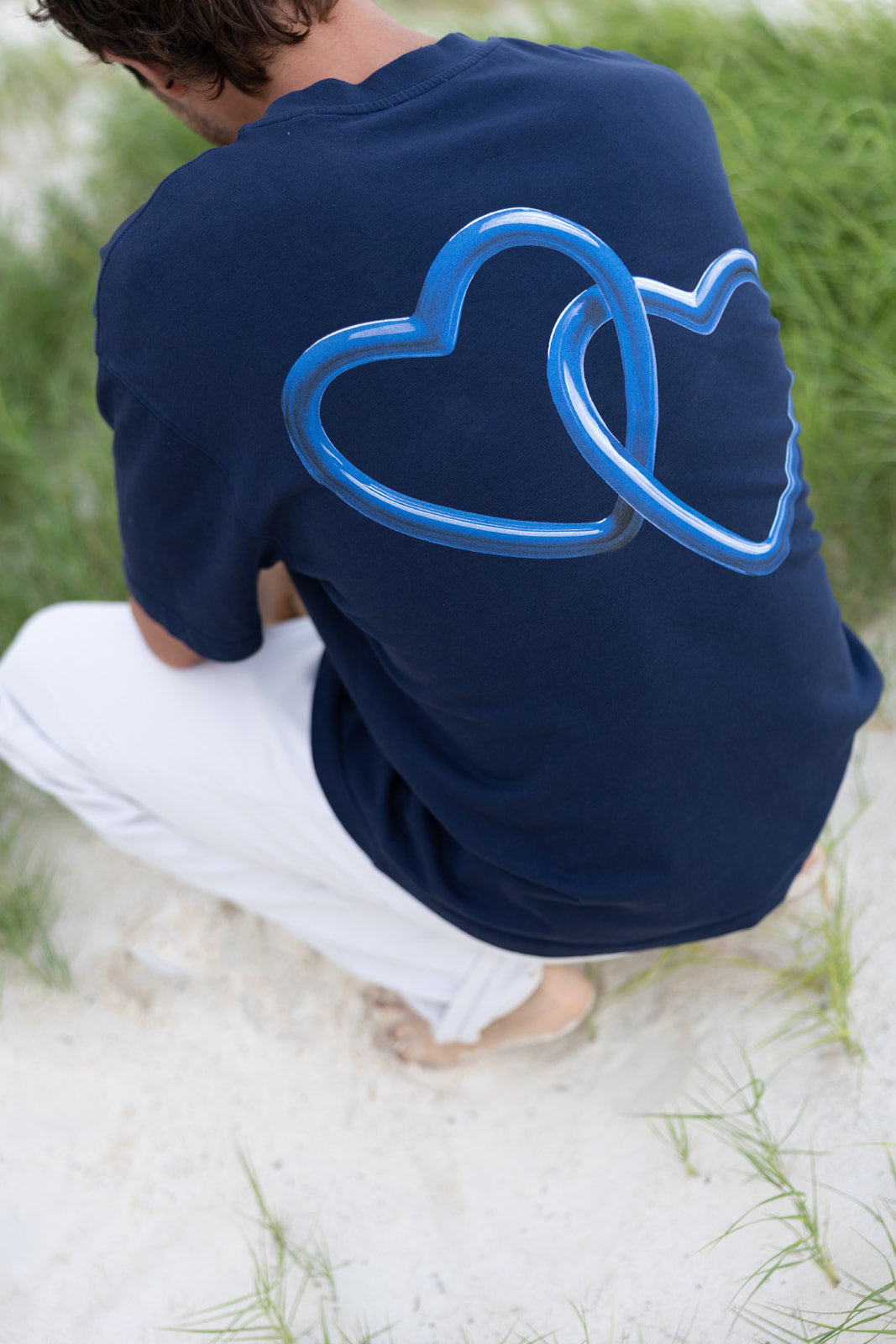 A person crouching outdoors on white sand, wearing a navy blue T-shirt with a large translucent blue interlocking heart graphic on the back and white pants.