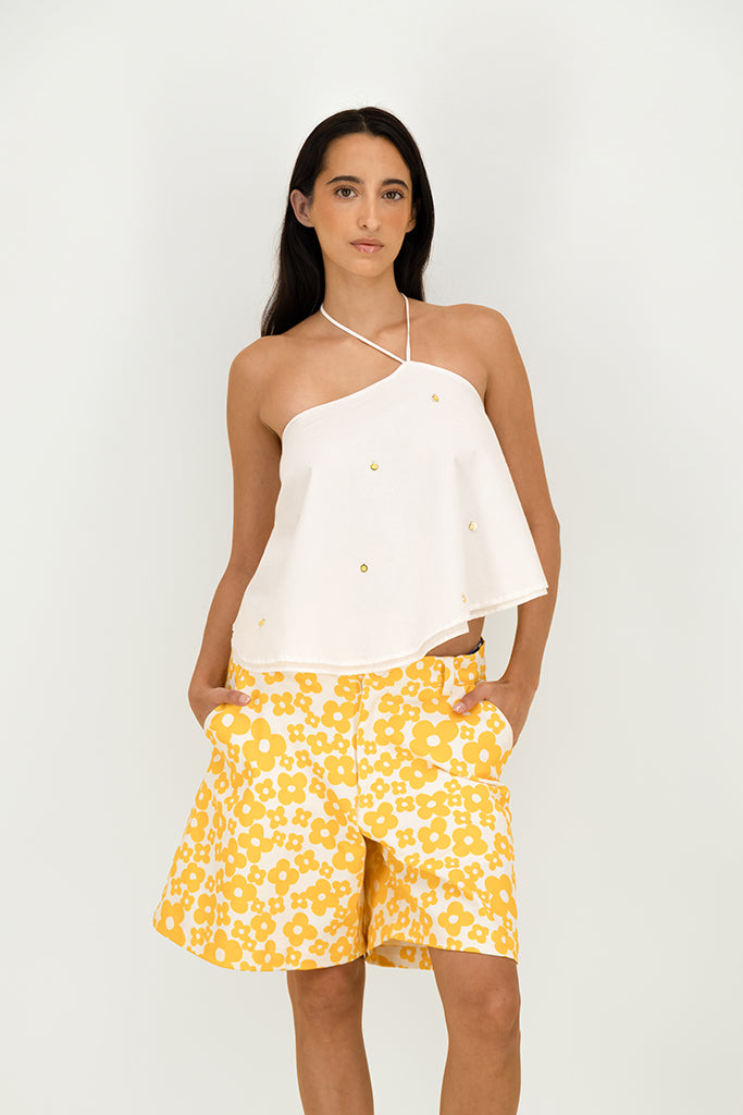 A woman modeling a white asymmetrical halter top with small gold button details and yellow floral print shorts against a light gray studio background.