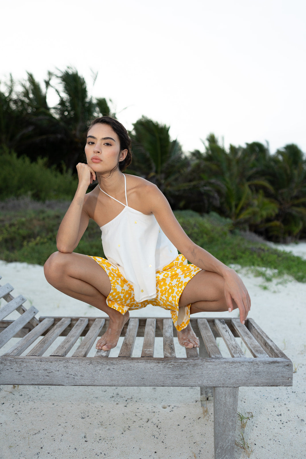 A woman crouching on a weathered wooden beach chair at the sand, wearing a white spaghetti-strap camisole and yellow floral shorts, with palm trees and white sand in the background.