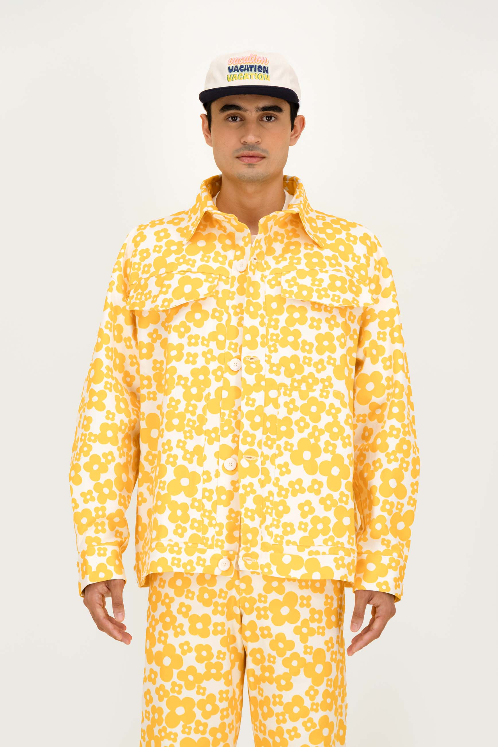 A man wearing a matching yellow and white floral print button-up shirt and pants set, paired with a white cap that reads 'MAISON COTON PARIS', photographed against a plain white studio background.