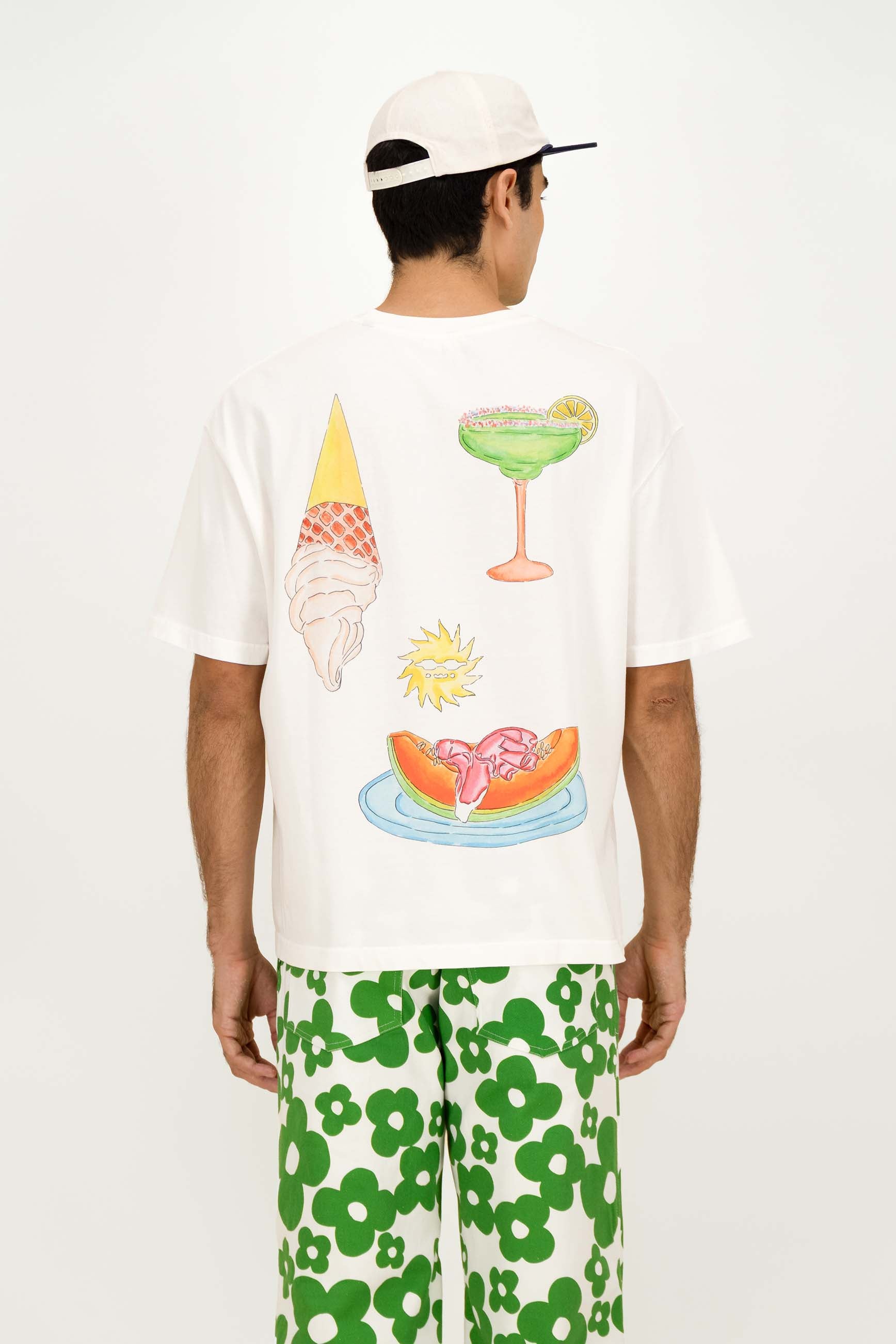 A man wearing a white t-shirt with a colorful back graphic of an ice cream cone, margarita, sun, and watermelon slice, paired with green and white floral shorts and a white cap.