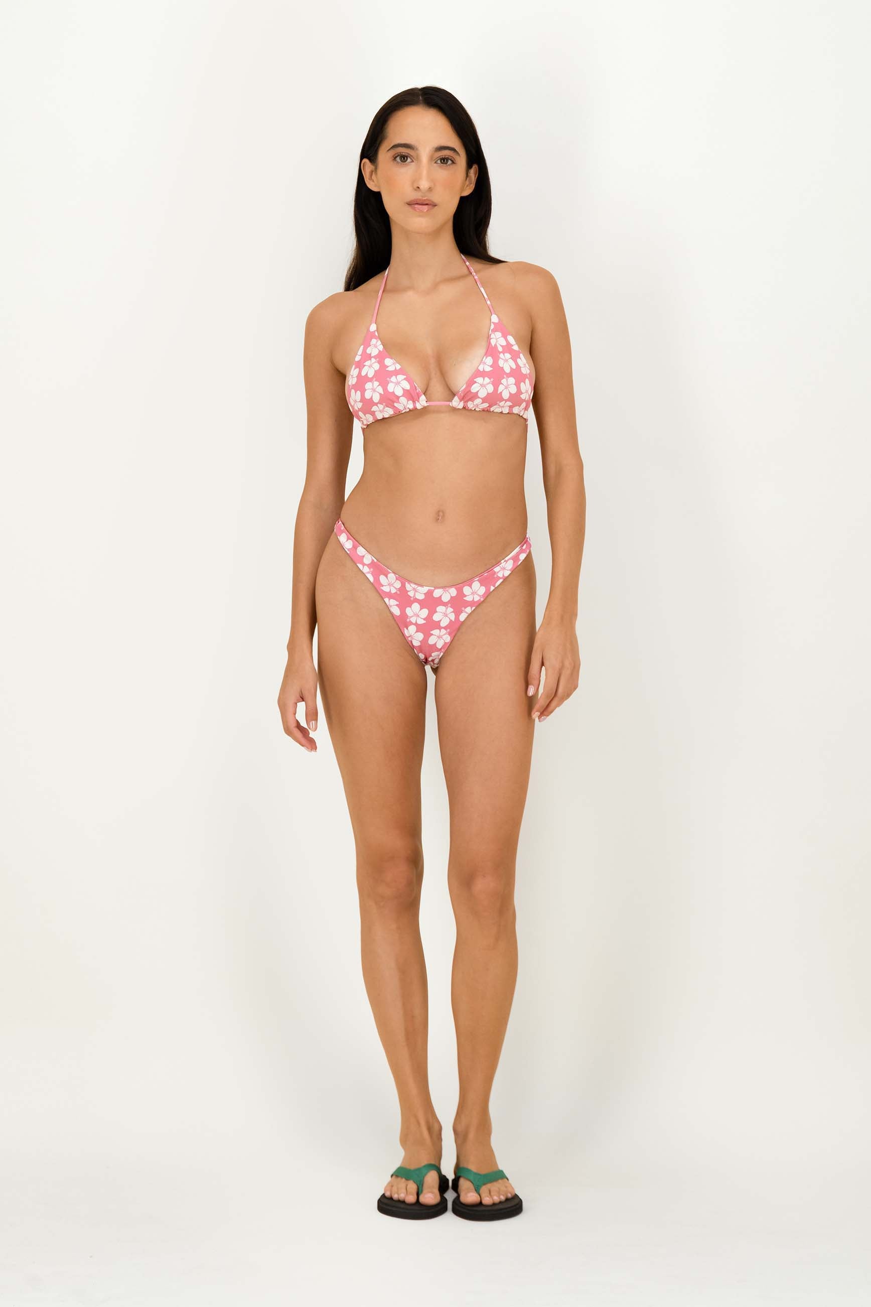A woman modeling a pink and white floral triangle bikini with green flip-flop sandals, standing against a plain white studio background.