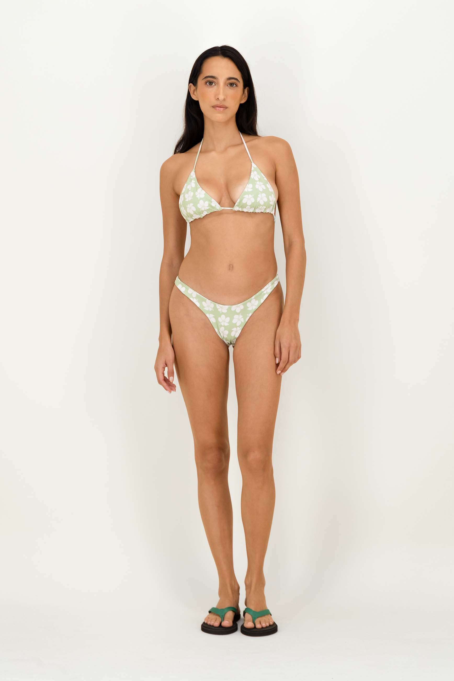A woman modeling a two-piece bikini with a light green and white floral pattern, paired with green and black flip-flops, against a plain white studio background.