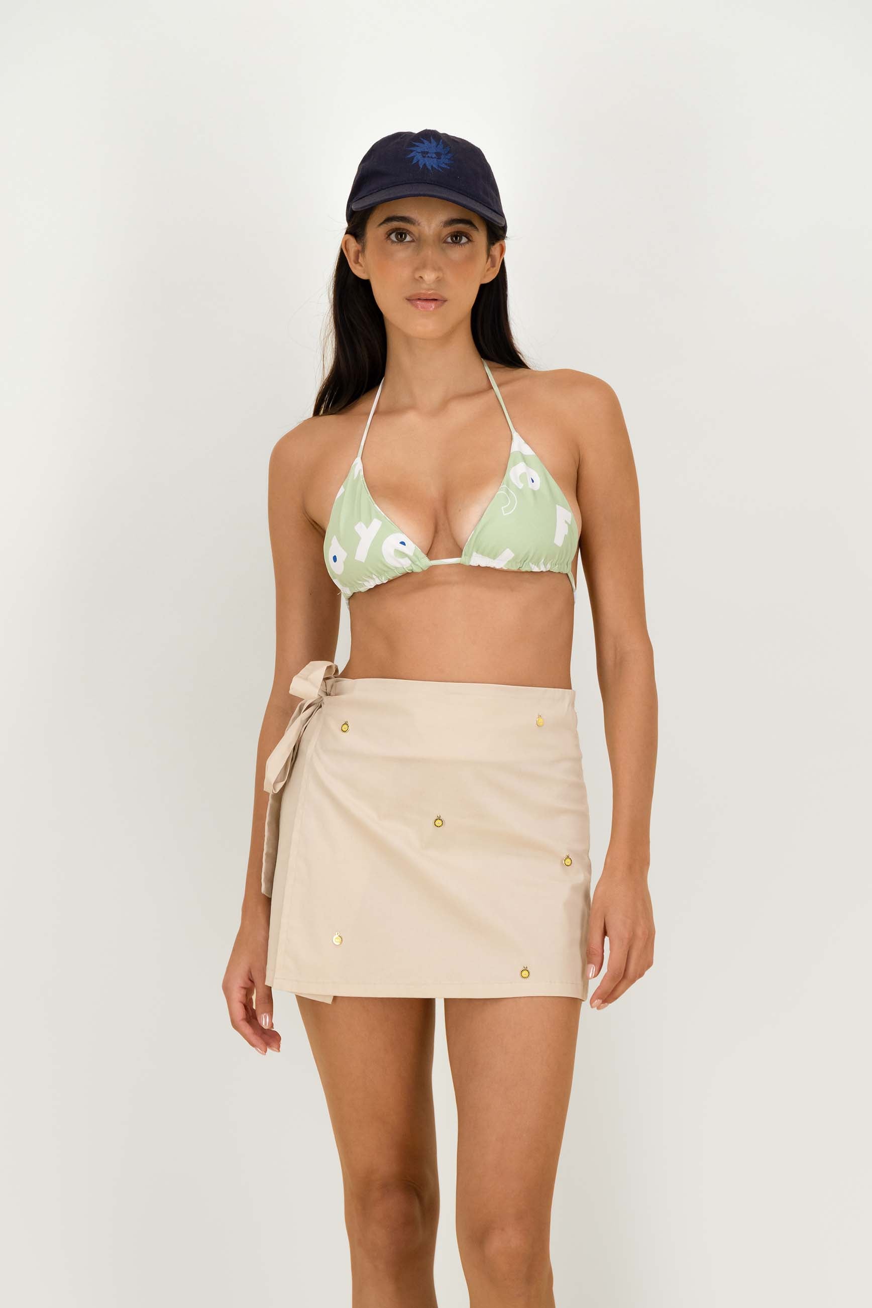 A model wearing a light green bikini top with a white floral pattern, a cream-colored wrap mini skirt with gold buttons, and a navy blue baseball cap, posed against a solid off-white studio background.