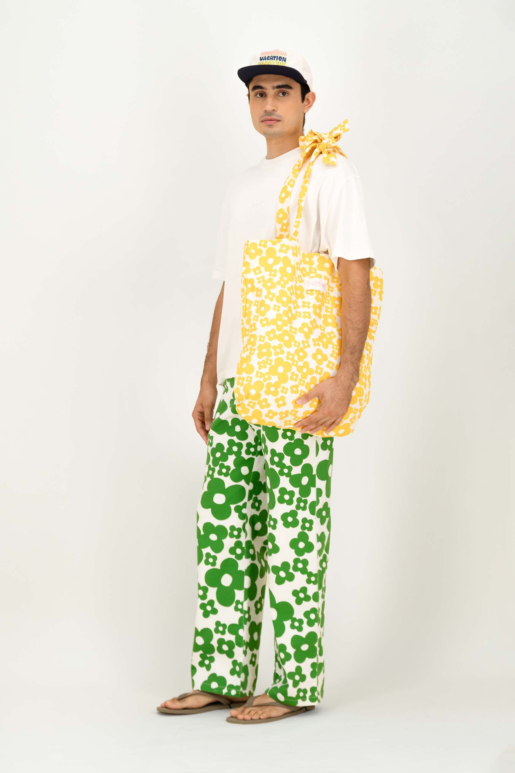 A man modeling a white t-shirt, green and white floral pants, a white cap, and carrying a yellow and white floral tote bag over his shoulder, set against a plain white studio background.