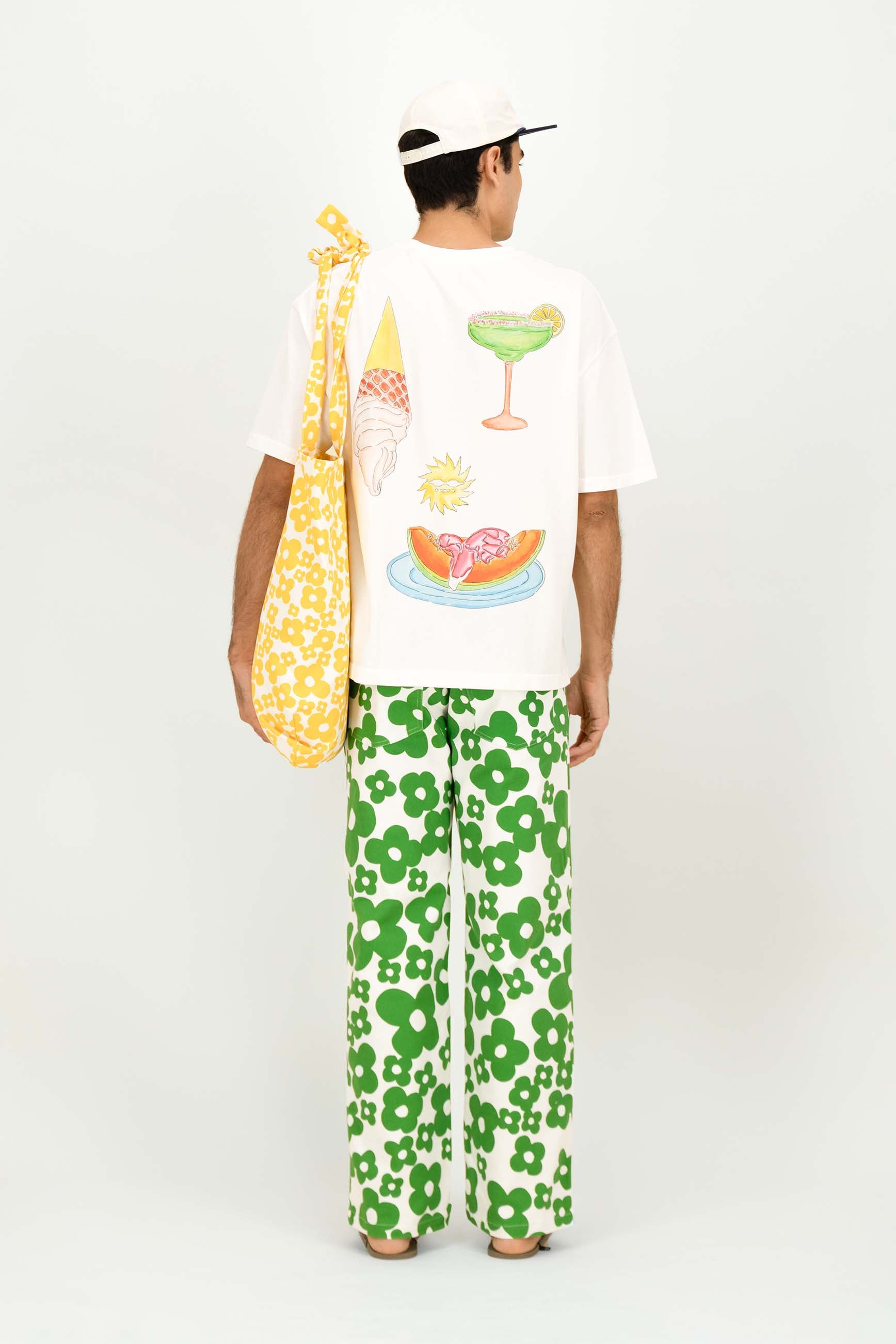 A model shown from the back wearing a white graphic t-shirt with colorful food illustrations, green and white patterned pants, a white cap, and carrying a yellow patterned shoulder bag against a plain white background.