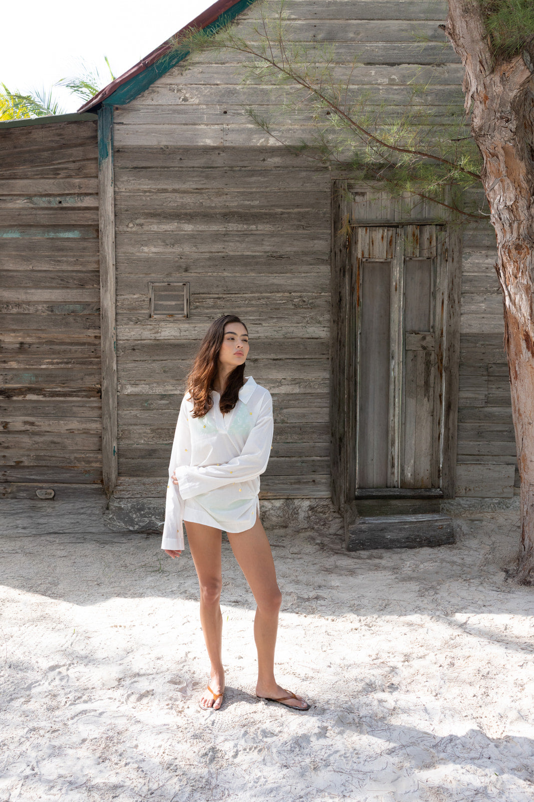 A woman stands barefoot on sandy ground in front of a rustic wooden hut, wearing a loose white long-sleeve transparent tunic