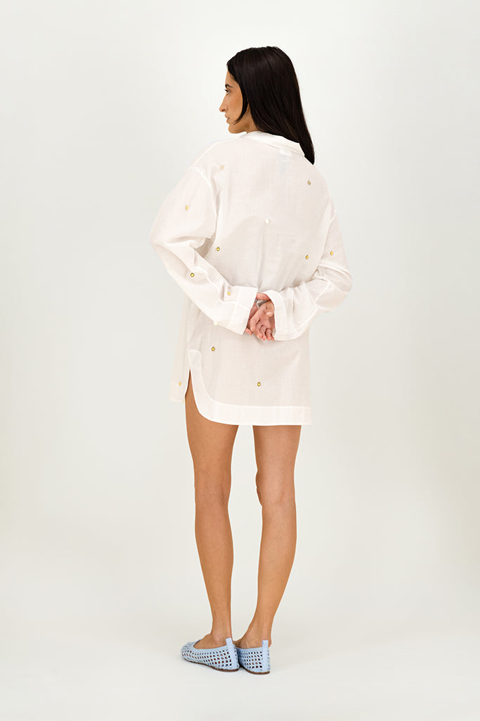 A woman shown from the back wearing a white long-sleeve button-up shirt dress with small gold embroidered details, paired with light blue and white checkered slip-on shoes, against a plain white studio background.