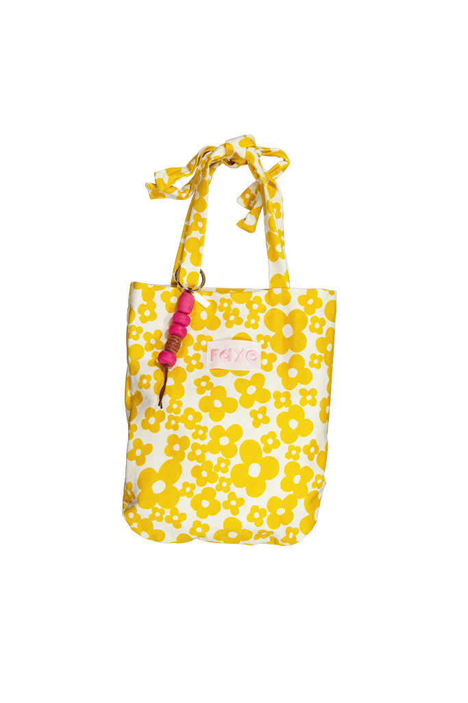 A yellow and white floral tote bag with matching fabric handles, a pink beaded keychain charm, and a small pink brand label on the front, shown against a plain white background.
