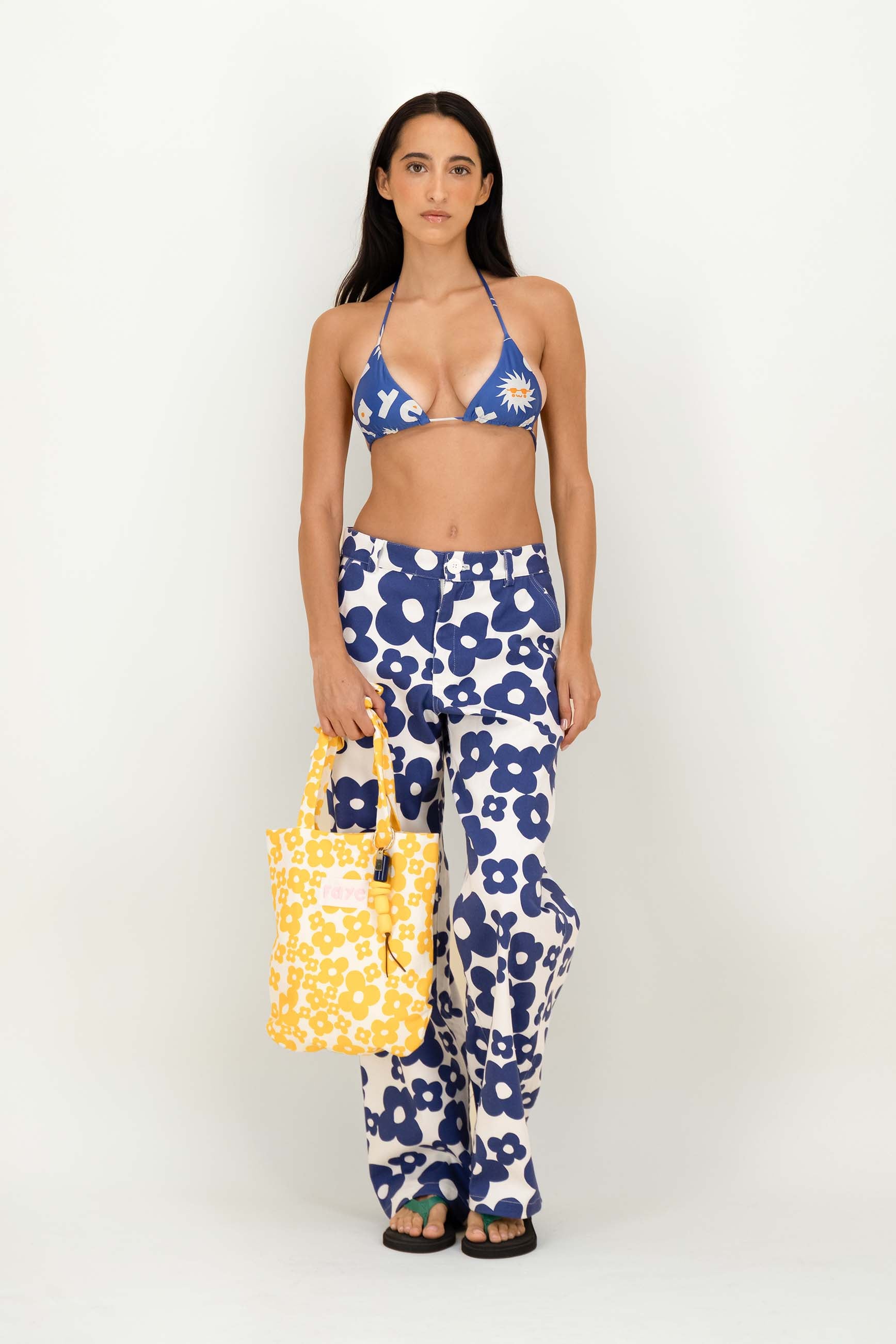 A woman stands against a plain white studio background wearing a matching two-piece swimsuit and pants set with a bold blue and white floral pattern, holding a yellow tote bag.