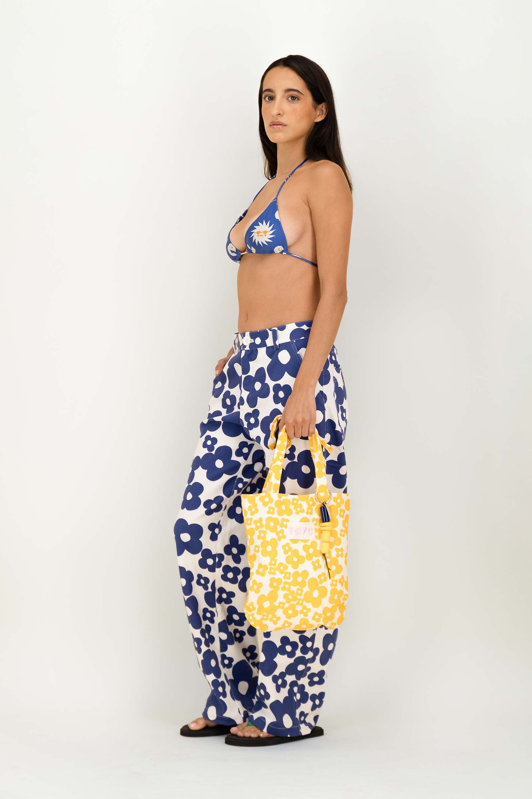 A woman stands in profile wearing a blue and white floral bikini top, matching wide-leg pants, and holding a yellow tote bag with a white floral print against a plain light gray studio background.