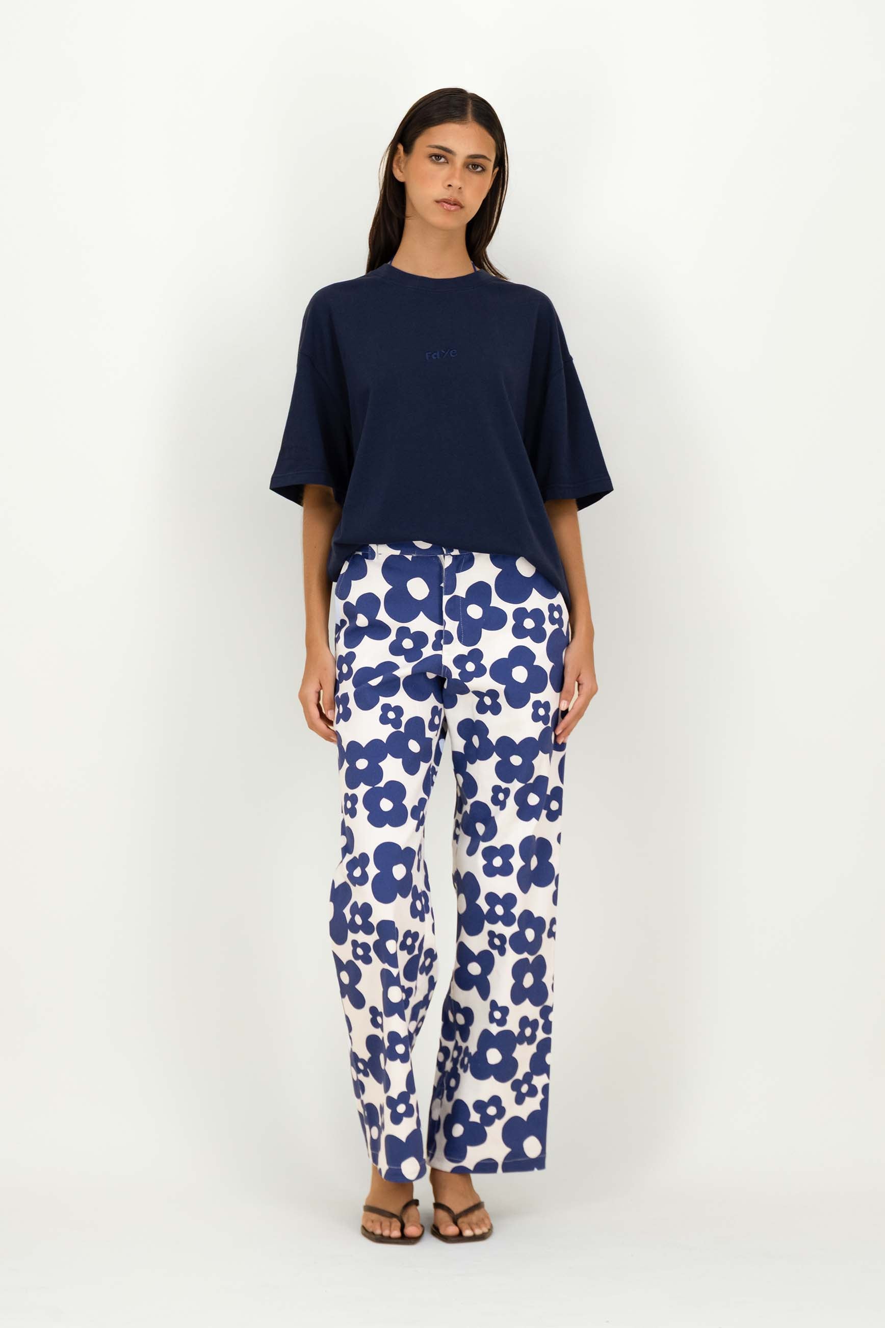 A woman modeling a navy blue short-sleeve top and white pants with a bold blue floral pattern, standing against a plain white studio background.