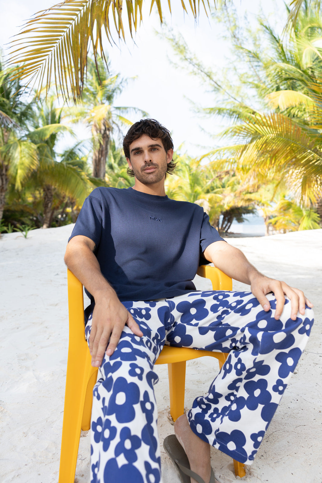 A man sitting on a yellow beach chair on white sand, wearing a navy blue t-shirt and blue-and-white floral print pants, with palm trees in the background.