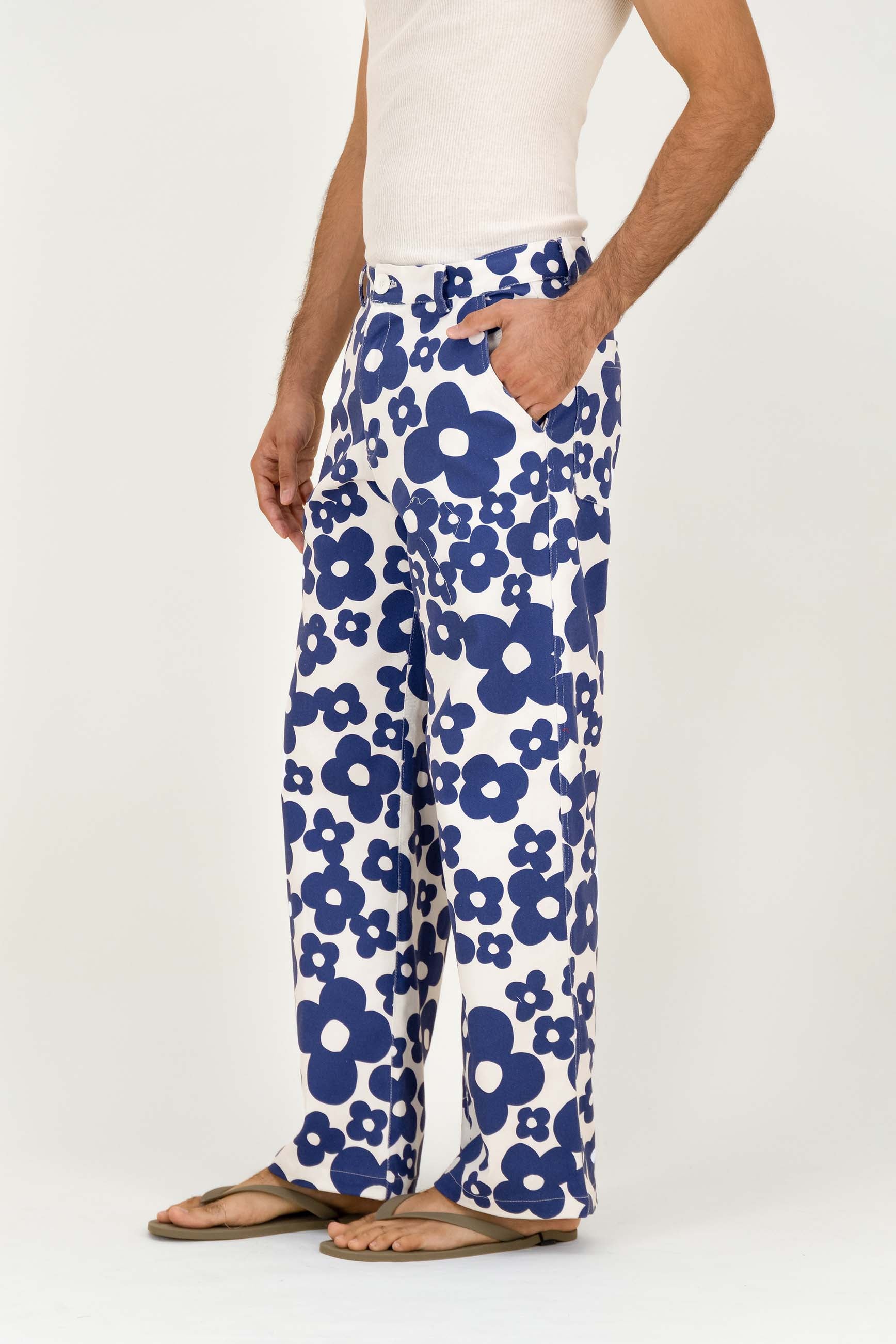 A person wearing white wide-leg pants with a bold blue floral pattern, paired with a white tank top and brown flip-flop sandals, shown against a plain light background.