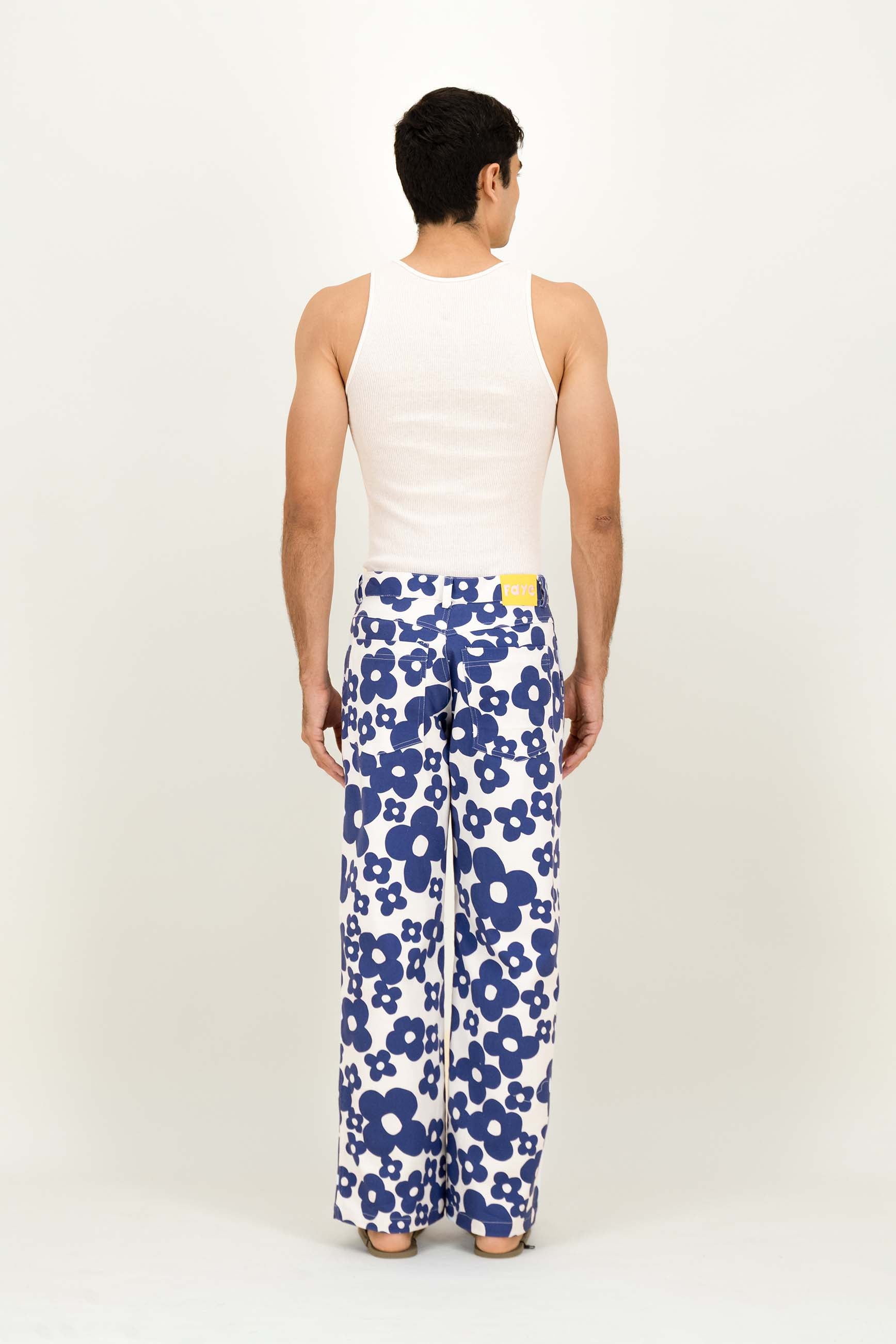 A man viewed from behind wearing a white ribbed tank top and white wide-leg trousers with a bold blue floral pattern, standing against a plain white studio background.