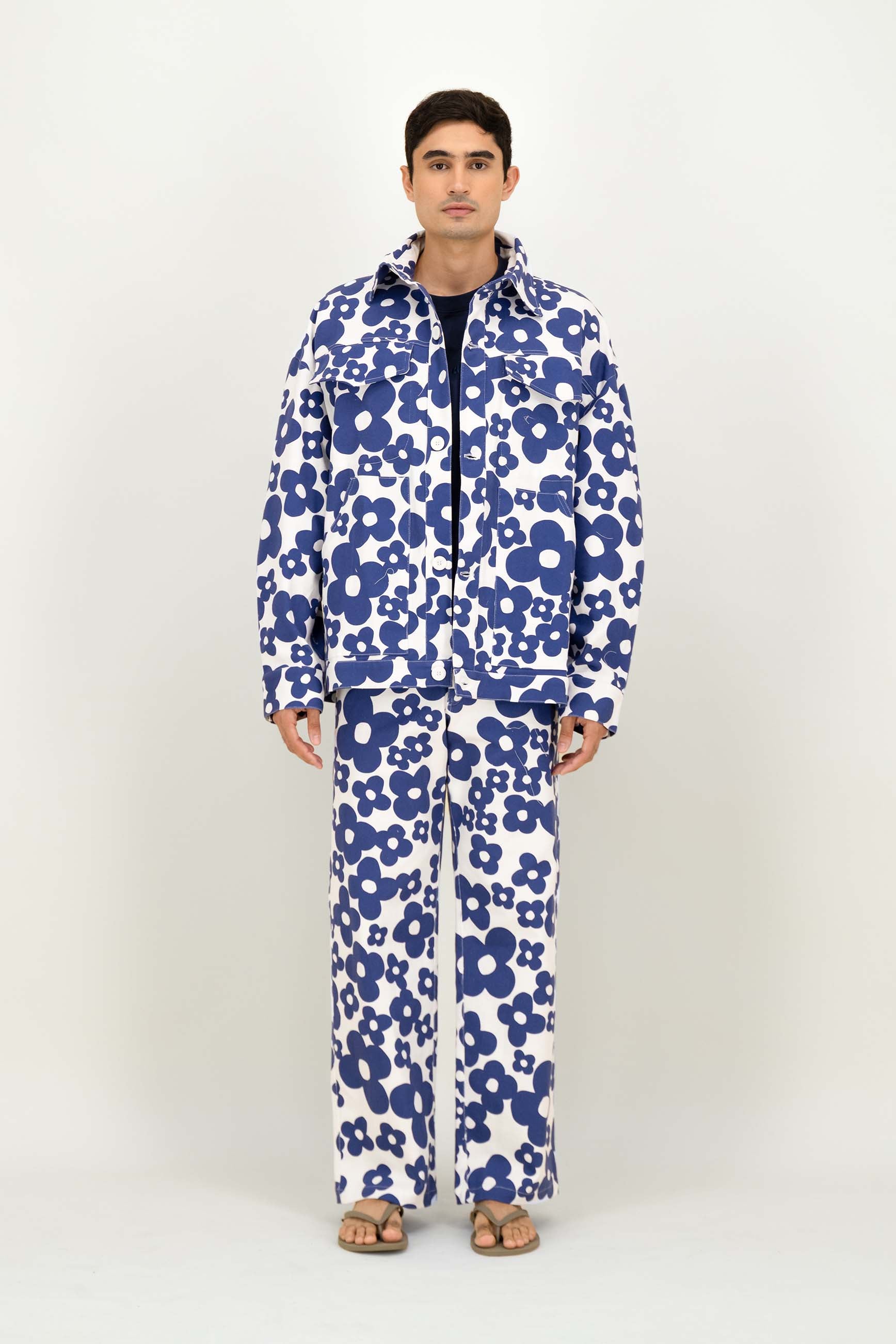 A man modeling a matching two-piece tracksuit with a bold blue and white floral pattern, featuring a zip-up jacket and wide-leg pants, shot against a plain light gray studio background.