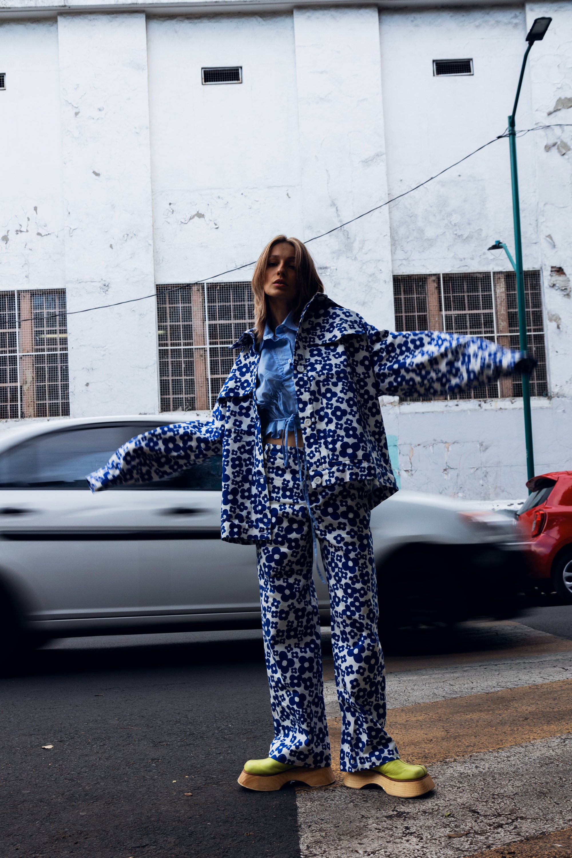 A model stands on a city street wearing a matching blue and white patterned jacket and trousers set with a light blue denim shirt underneath and bright yellow-green platform shoes. A white building and passing cars are in the background.