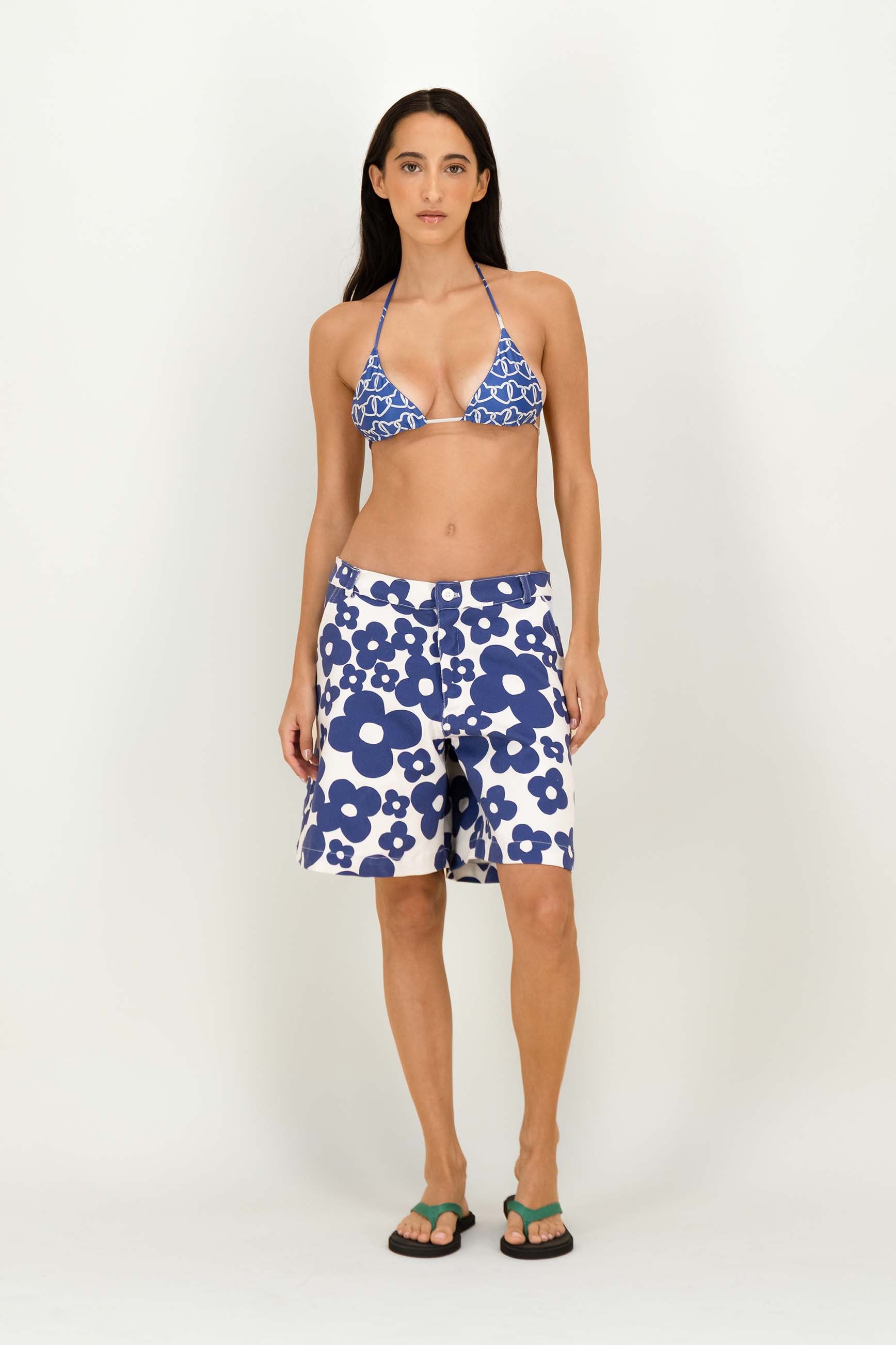 A woman modeling a two-piece swimwear set featuring a blue and white floral triangle bikini top and matching floral board shorts, paired with green flip-flops, against a plain light gray studio background.