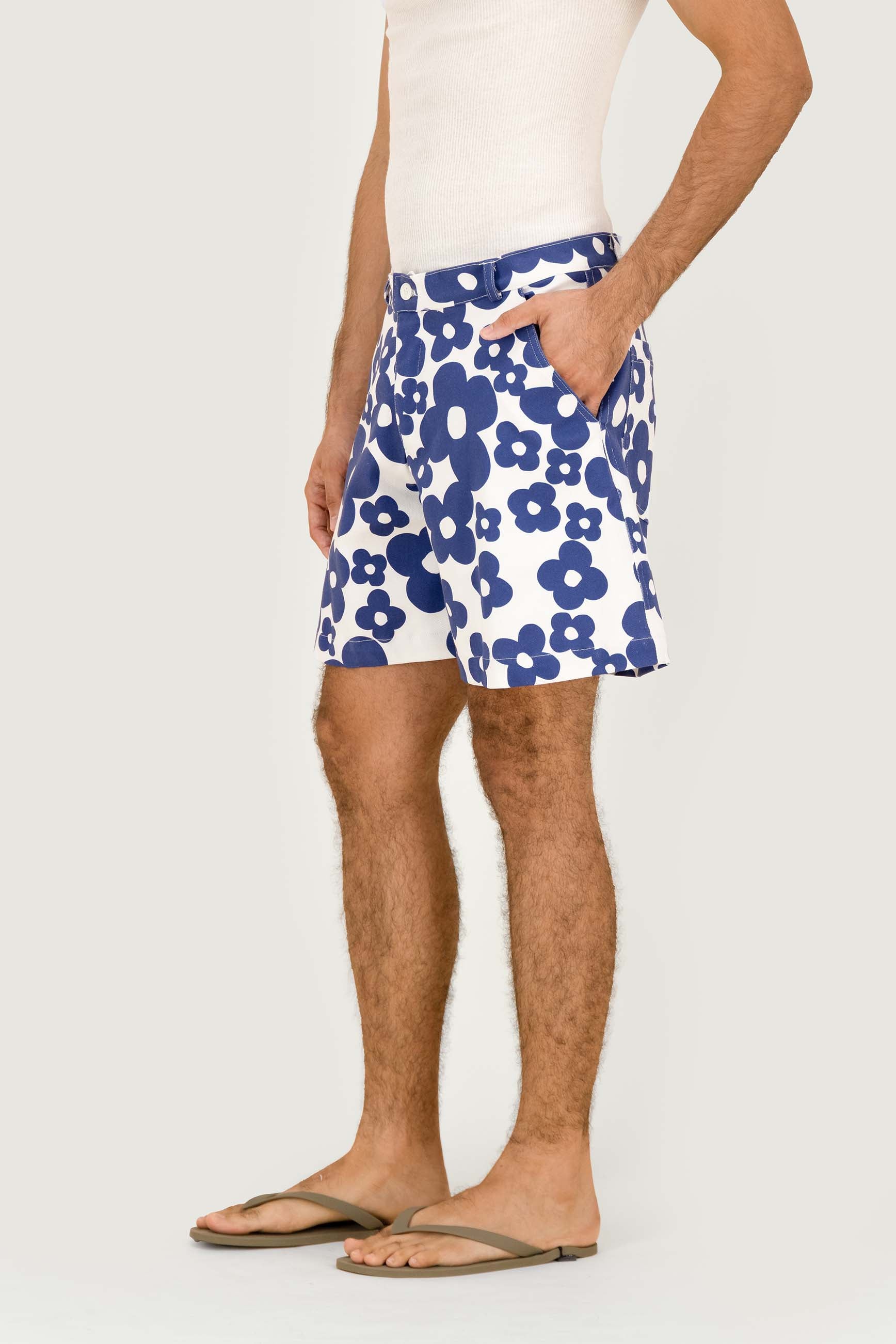 A man wearing blue and white floral print shorts, a white tank top, and brown flip-flops, shown from the waist down against a light gray studio background.
