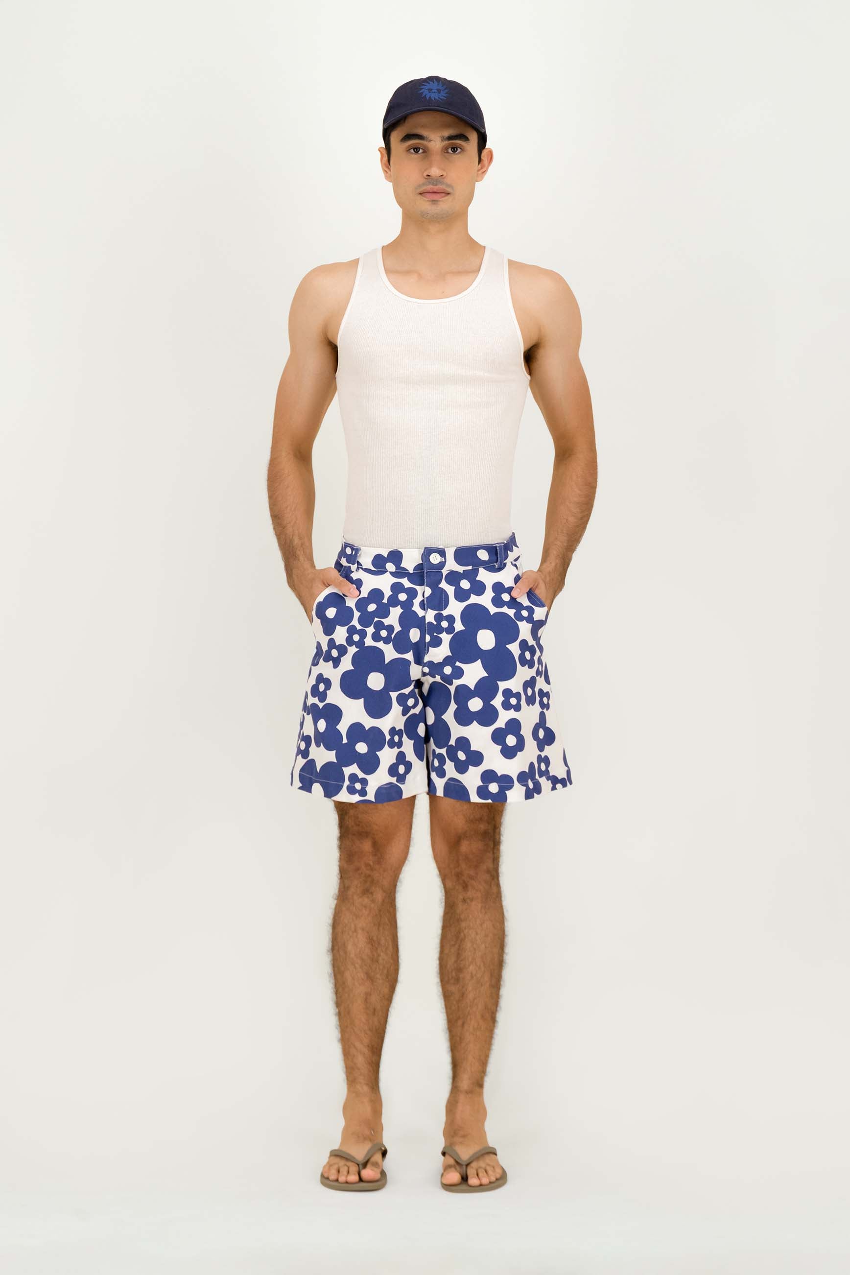 A man stands against a plain light background wearing a white tank top, blue and white floral shorts, a dark blue cap, and sandals.