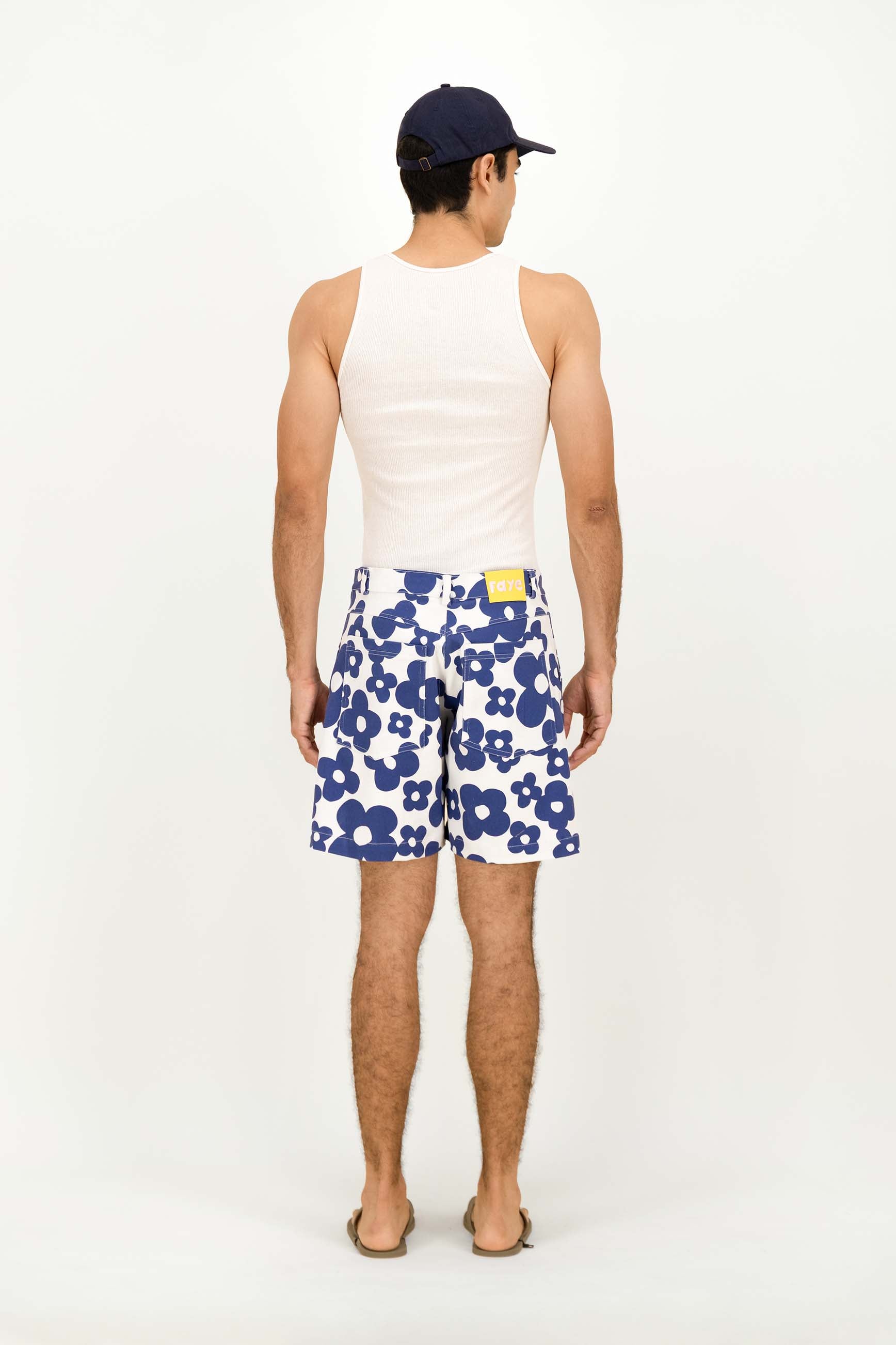 A man shown from the back wearing a white tank top, navy blue baseball cap, and white swim trunks with a bold blue floral pattern. He is standing against a plain white studio background.