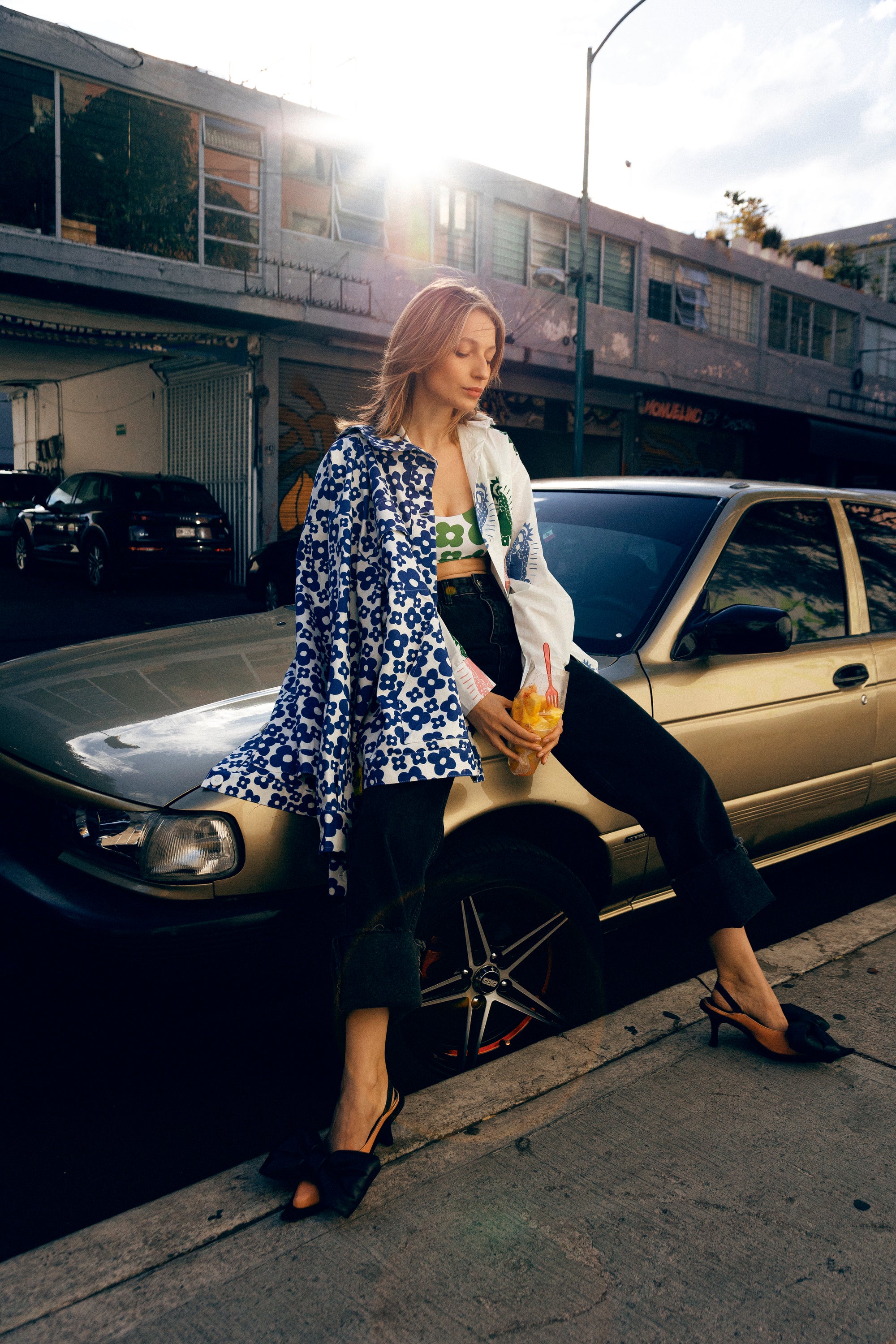 A woman posing outdoors next to a gold car on a city street, wearing a blue and white patterned long-sleeve vest, black pants, and black heels.