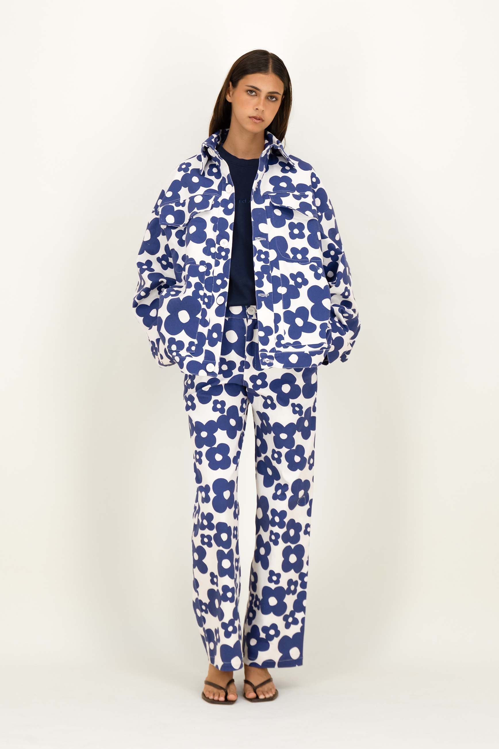 A model stands against a plain off-white studio background wearing a matching blue and white floral print vest and trousers set, styled with a dark navy top and brown sandals.