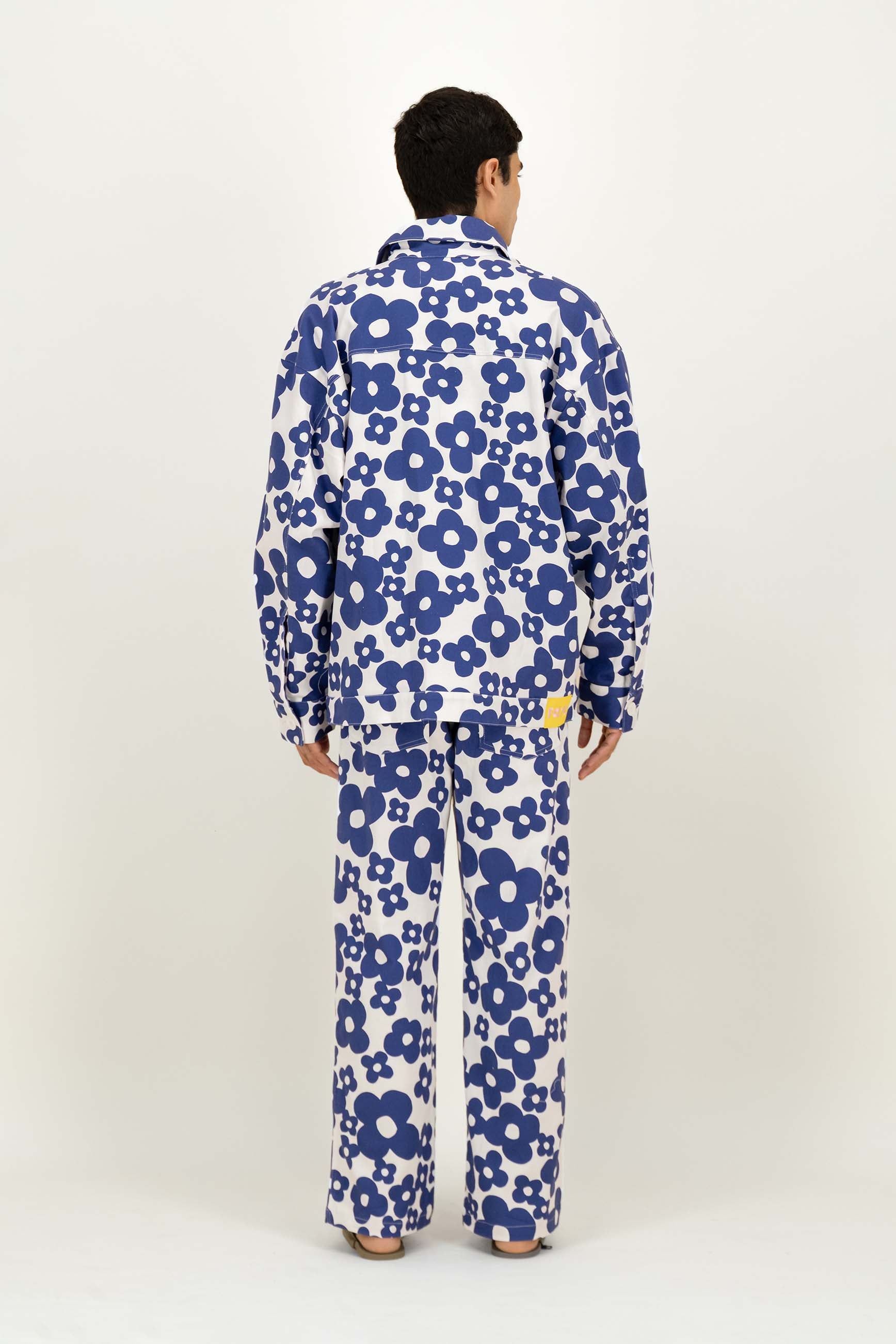 A man viewed from behind wearing a matching long-sleeve button-up vest and trousers set in a white fabric with a bold blue floral pattern, photographed against a plain off-white studio background.
