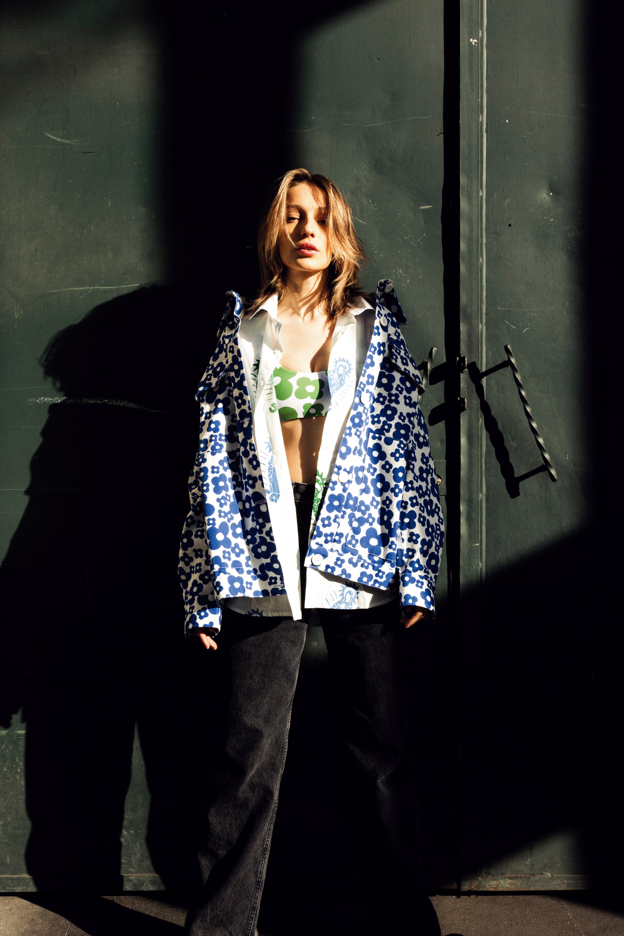 A model stands against a dark green door in dramatic, high-contrast sunlight and shadow, wearing a blue and white patterned open jacket over a green graphic crop top and dark wide-leg pants.