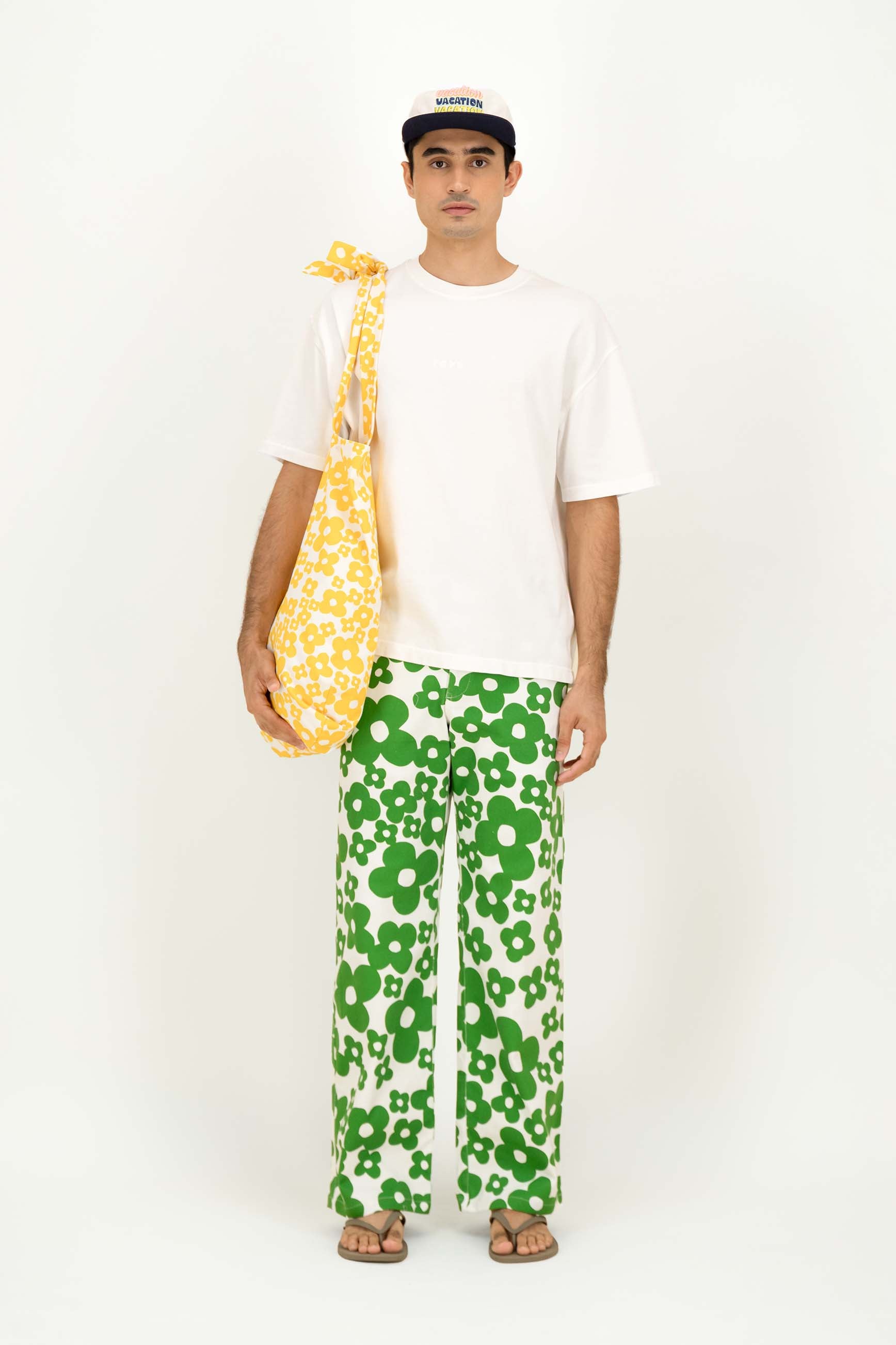 A man stands against a plain white studio background wearing a white T-shirt, green-and-white floral wide-leg pants, and a black-and-white cap. He is holding a yellow-and-white patterned shoulder bag.