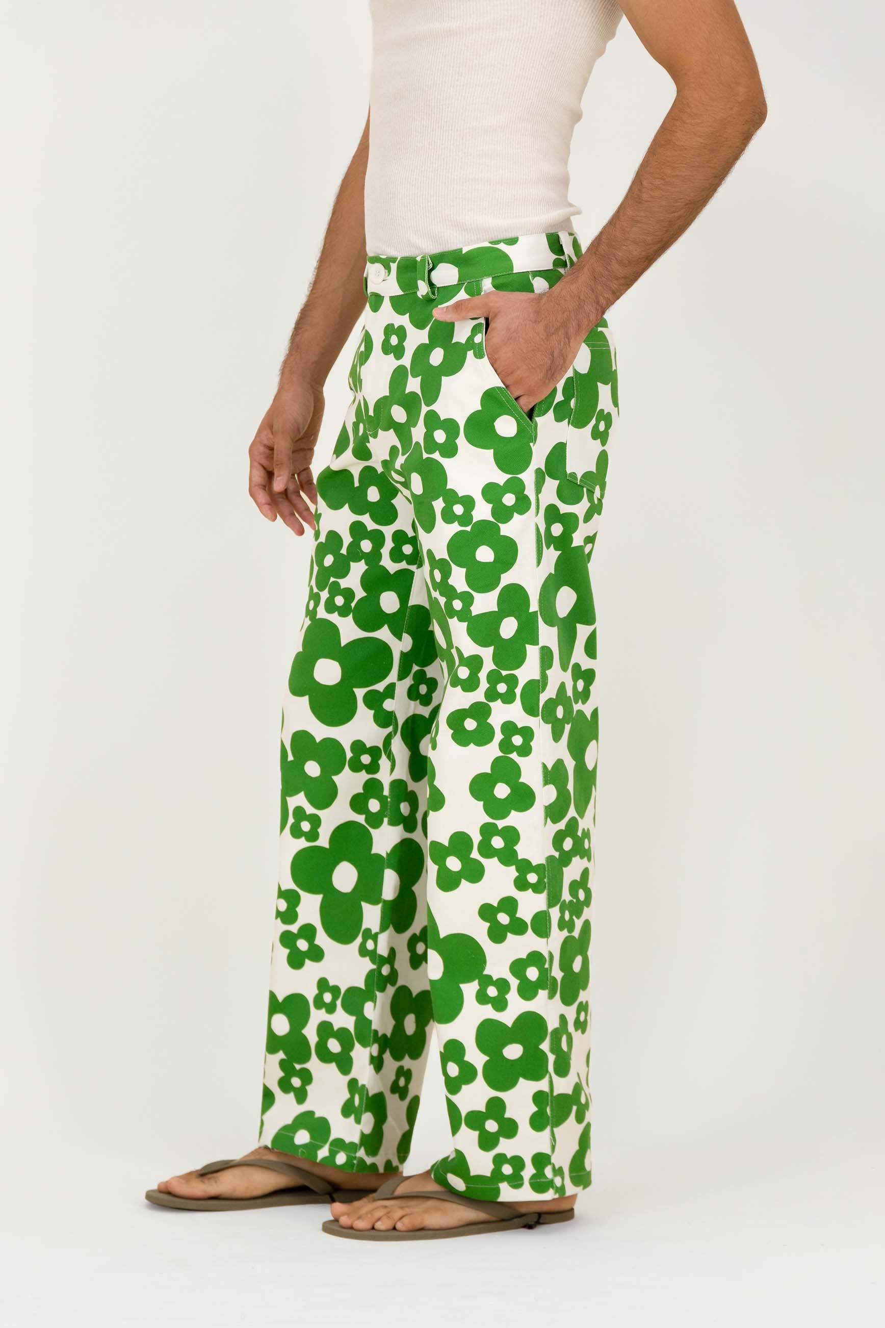 A person wearing wide-leg white pants with a bold green floral pattern, styled with a white tank top and tan sandals against a plain white studio background.