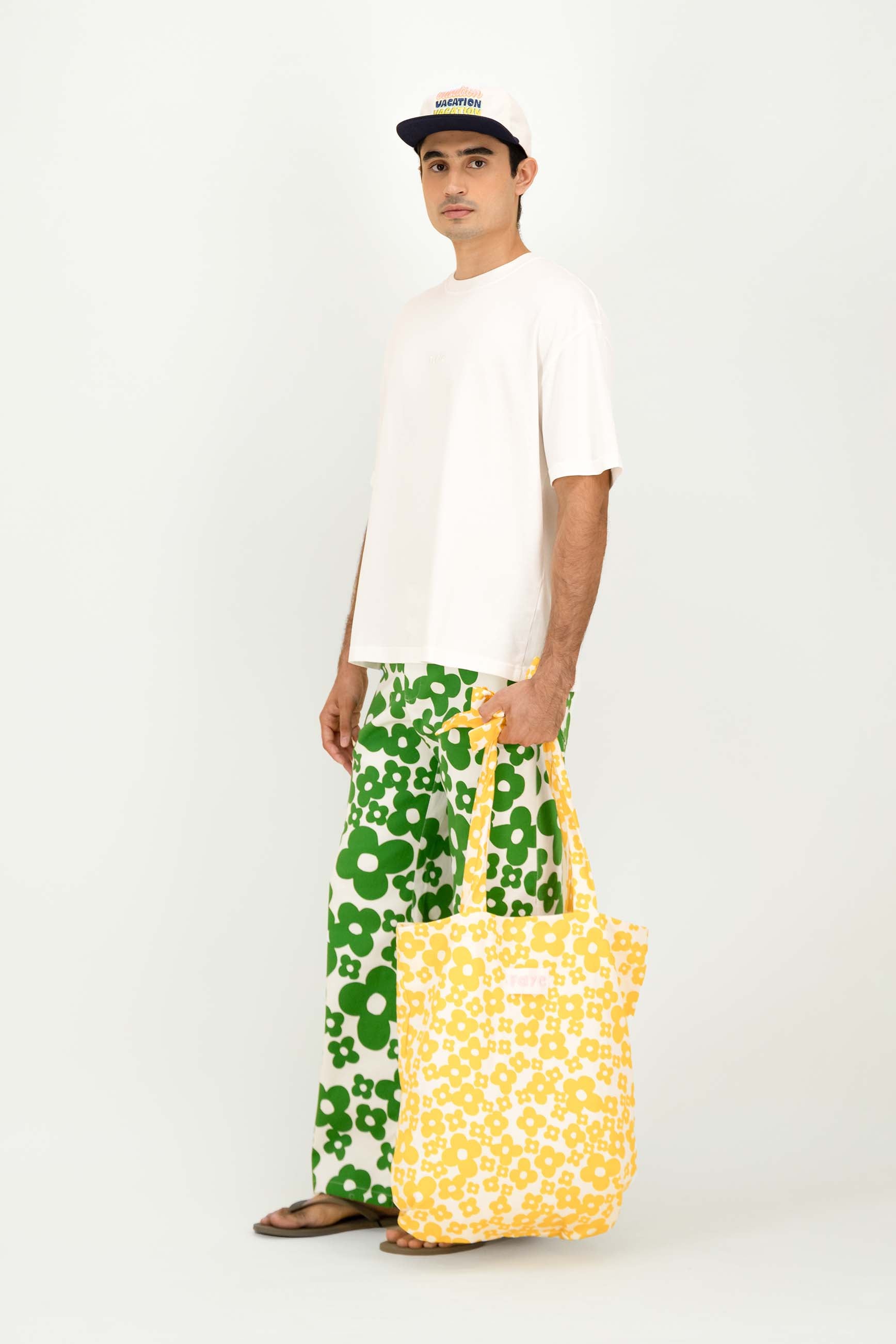 A man wearing a white t-shirt, green and white floral pants, and a black and white cap stands against a white studio background, holding a yellow and white floral tote bag.
