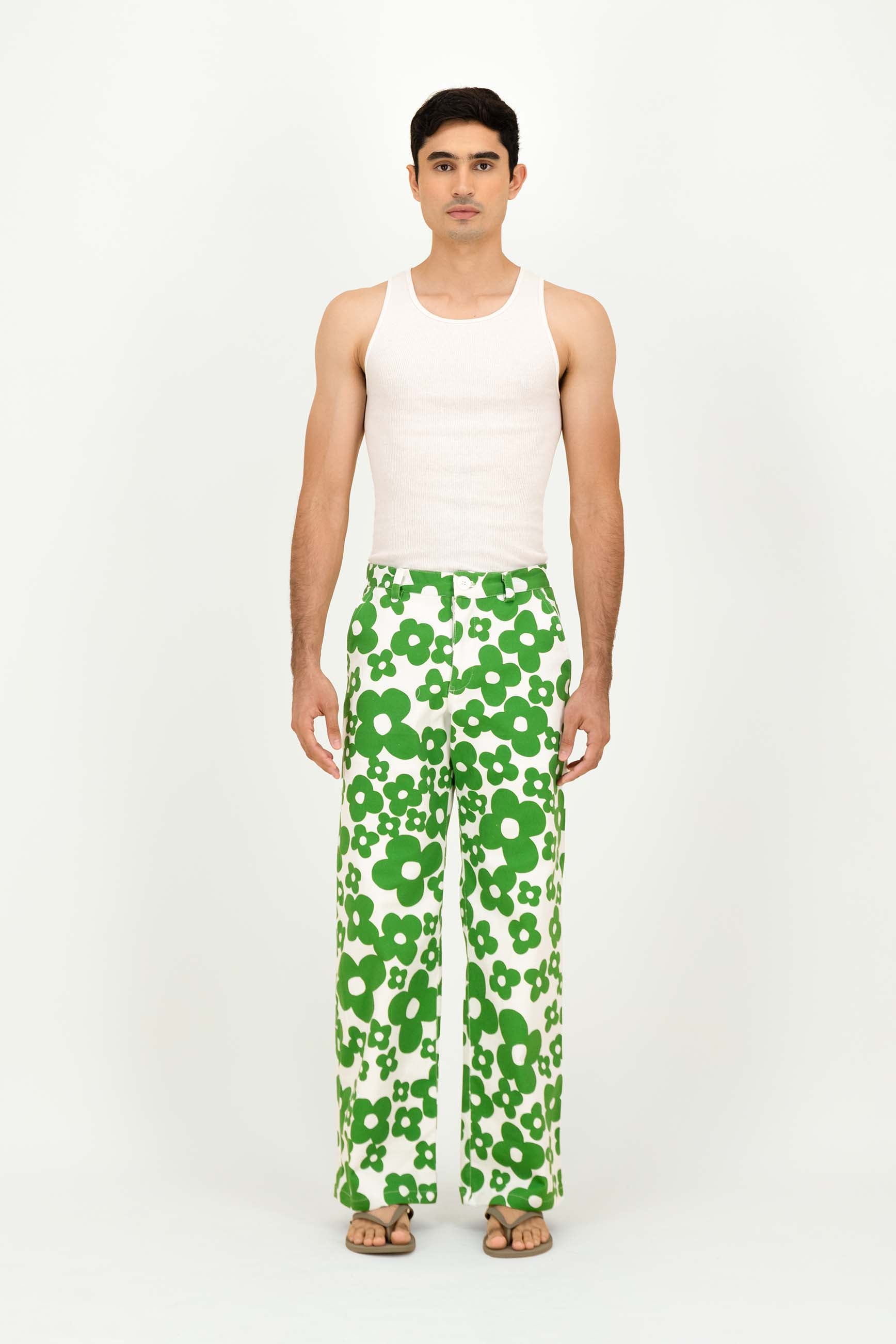 A man stands against a plain white studio background wearing a white ribbed tank top and white wide-leg pants with a bold green floral pattern. He is wearing tan sandals.