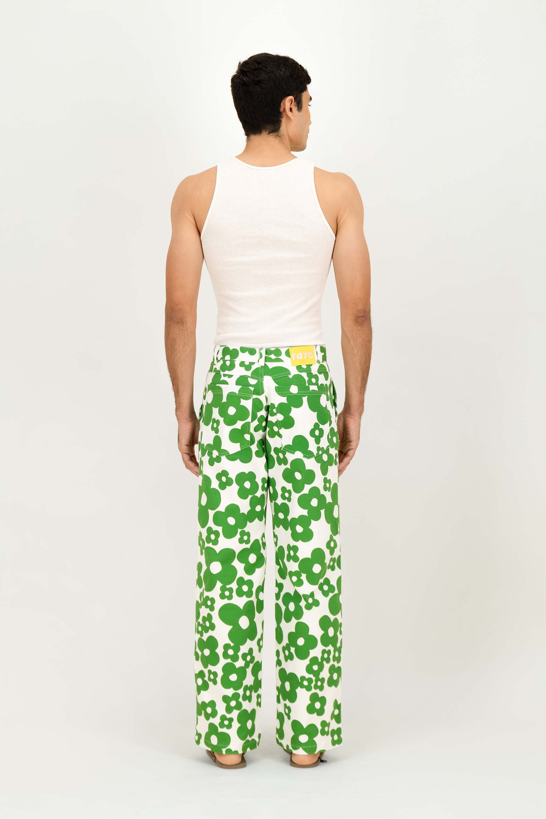 A man viewed from behind wearing white wide-leg trousers with a bold green floral pattern, a white tank top, and glasses, standing against a plain white studio background.