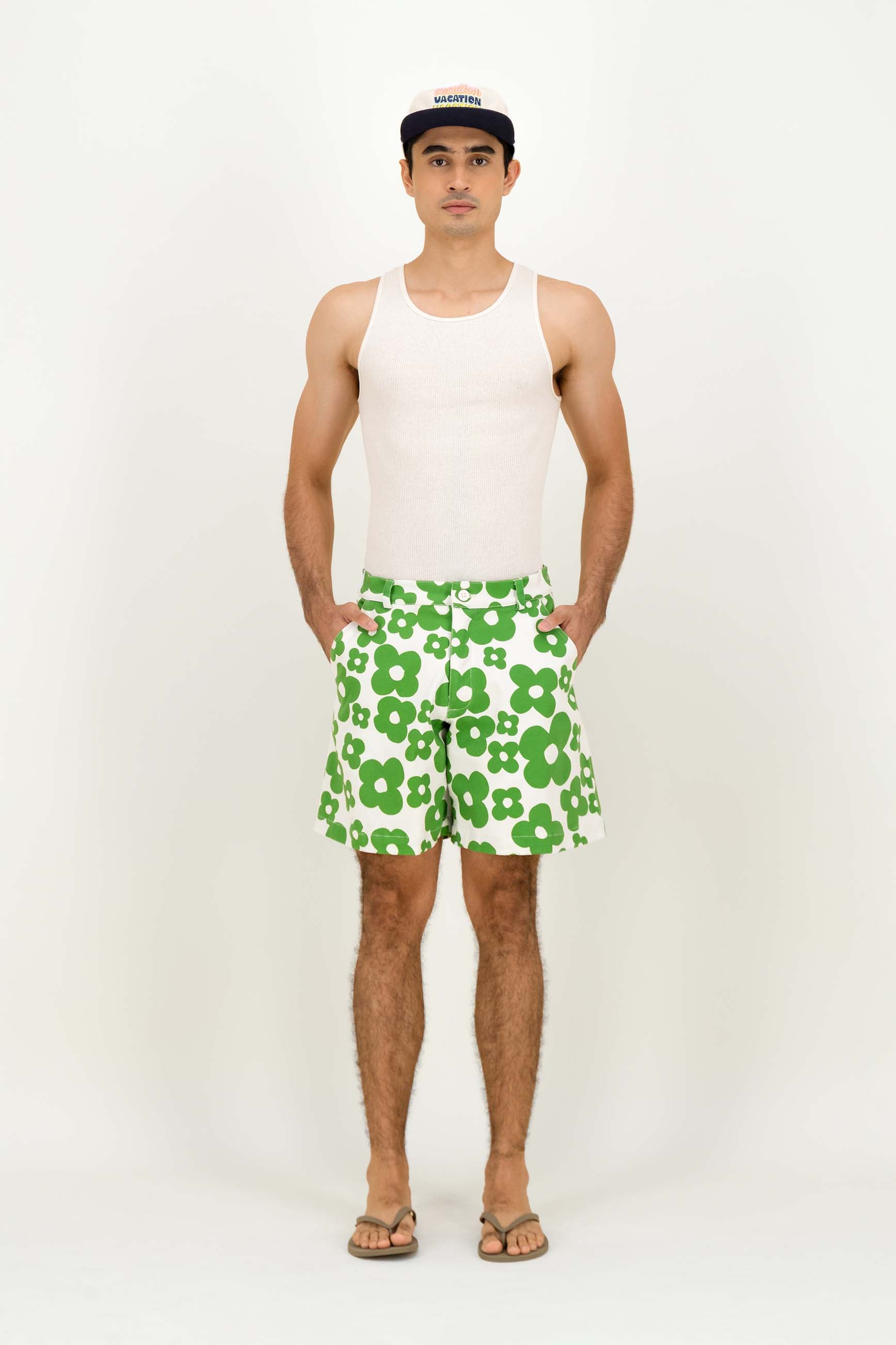 A man modeling a white tank top and green-and-white floral print shorts against a plain white studio background.