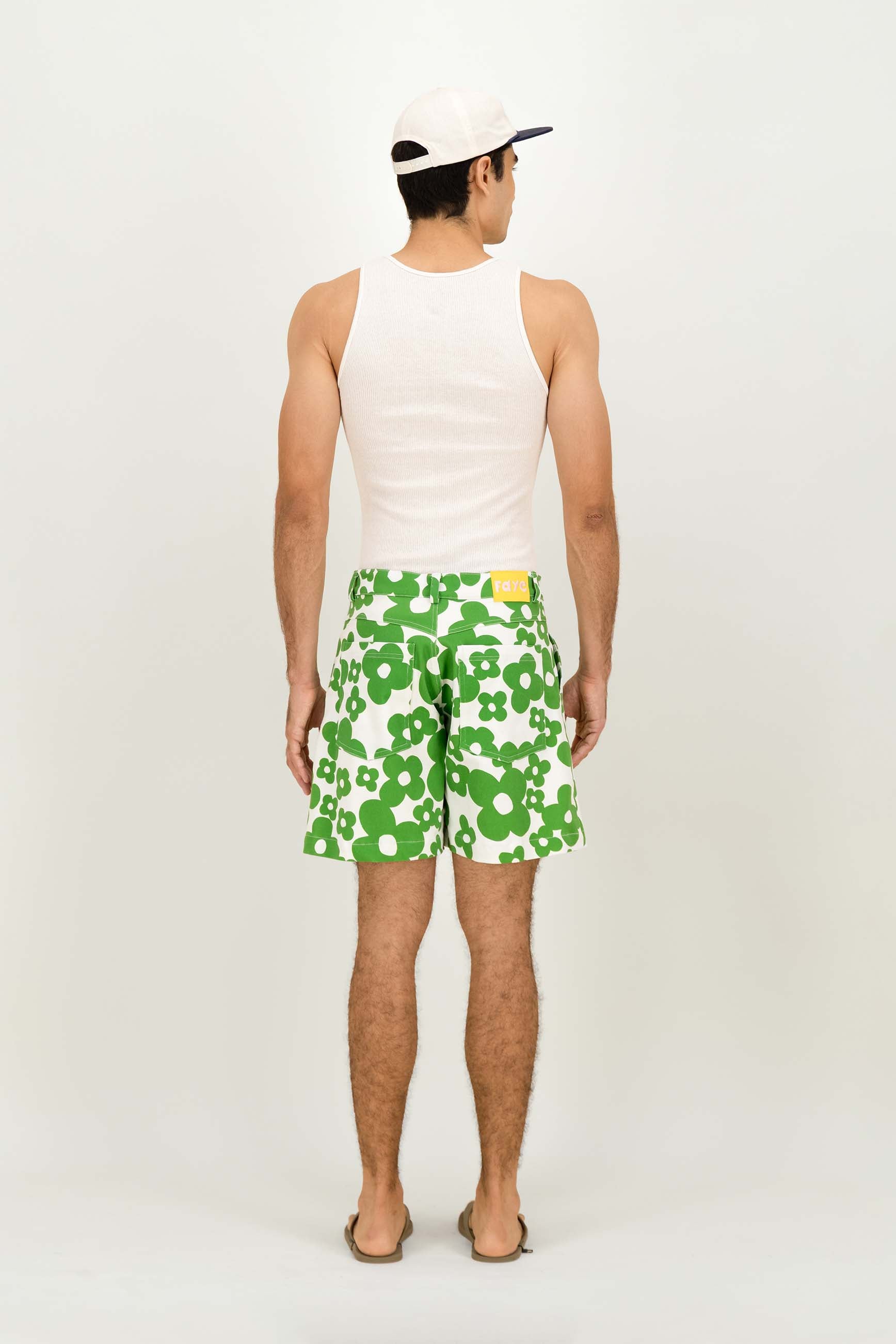 A man viewed from behind wearing green and white patterned swim trunks, a white tank top, and a white baseball cap against a plain light gray studio background.