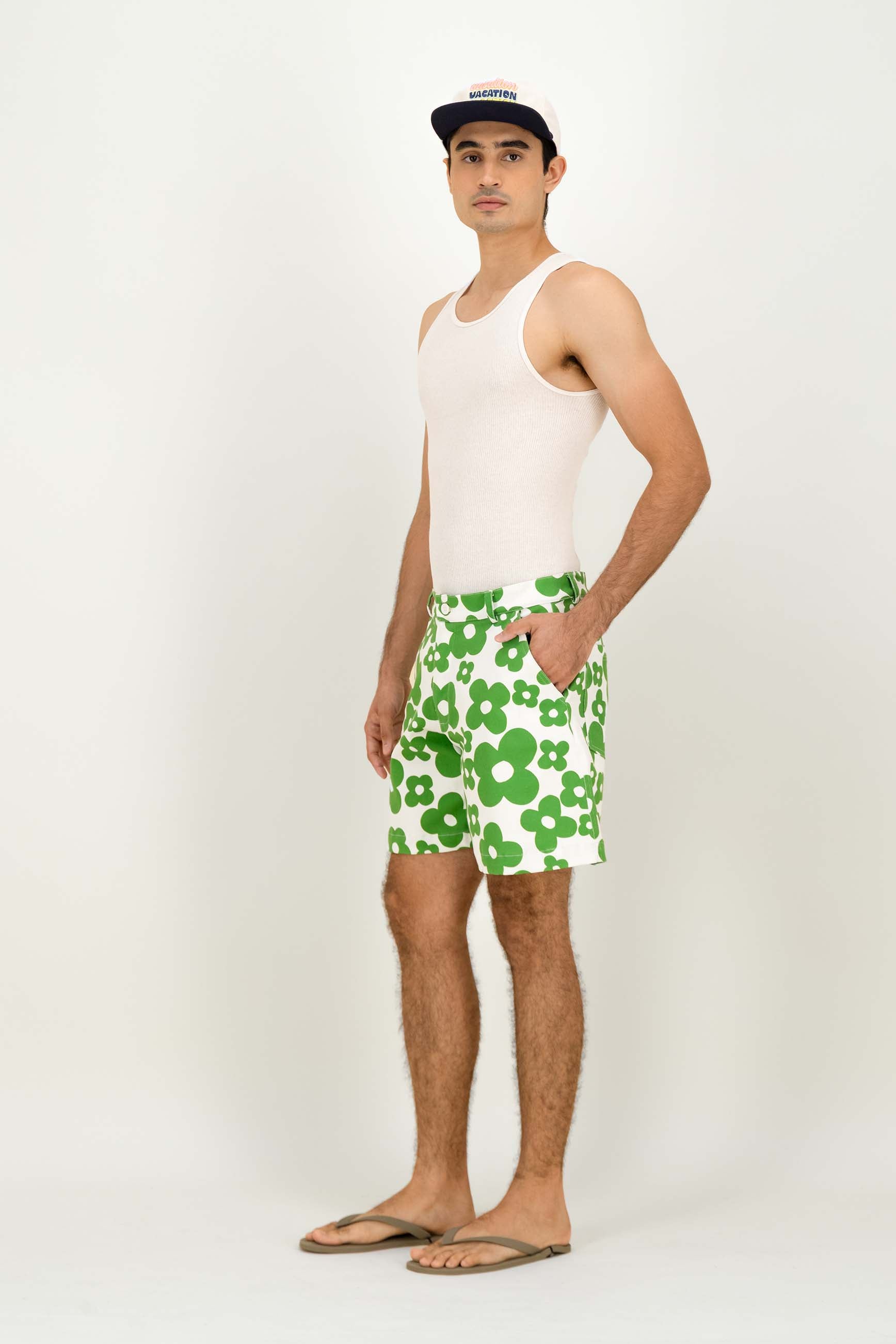A man modeling green and white clover-patterned shorts, a white tank top, and a black baseball cap against a plain white studio background.