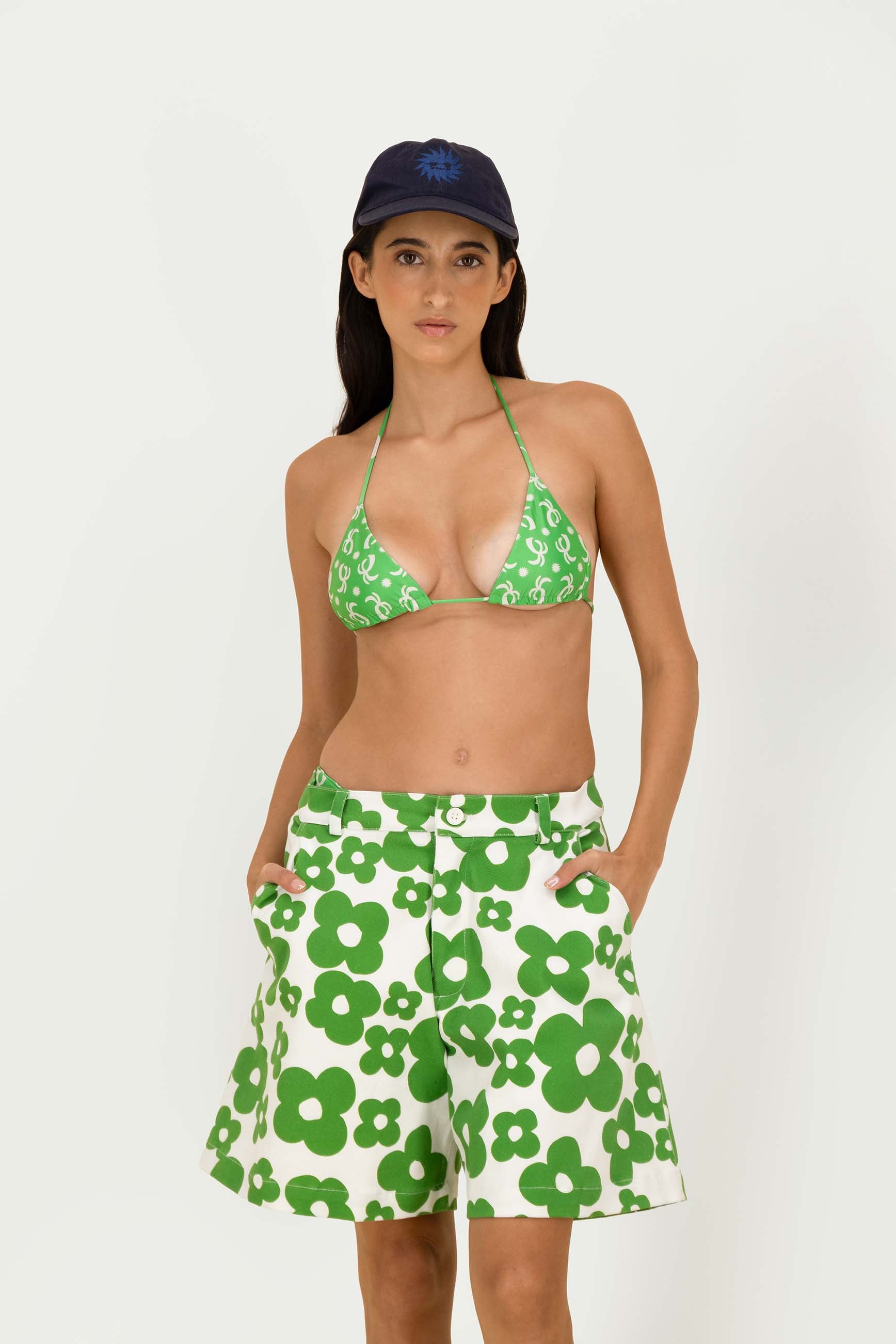 A woman modeling a green and white floral print two-piece swimwear set, consisting of a triangle bikini top and matching shorts, paired with a navy blue baseball cap, against a plain white studio background.